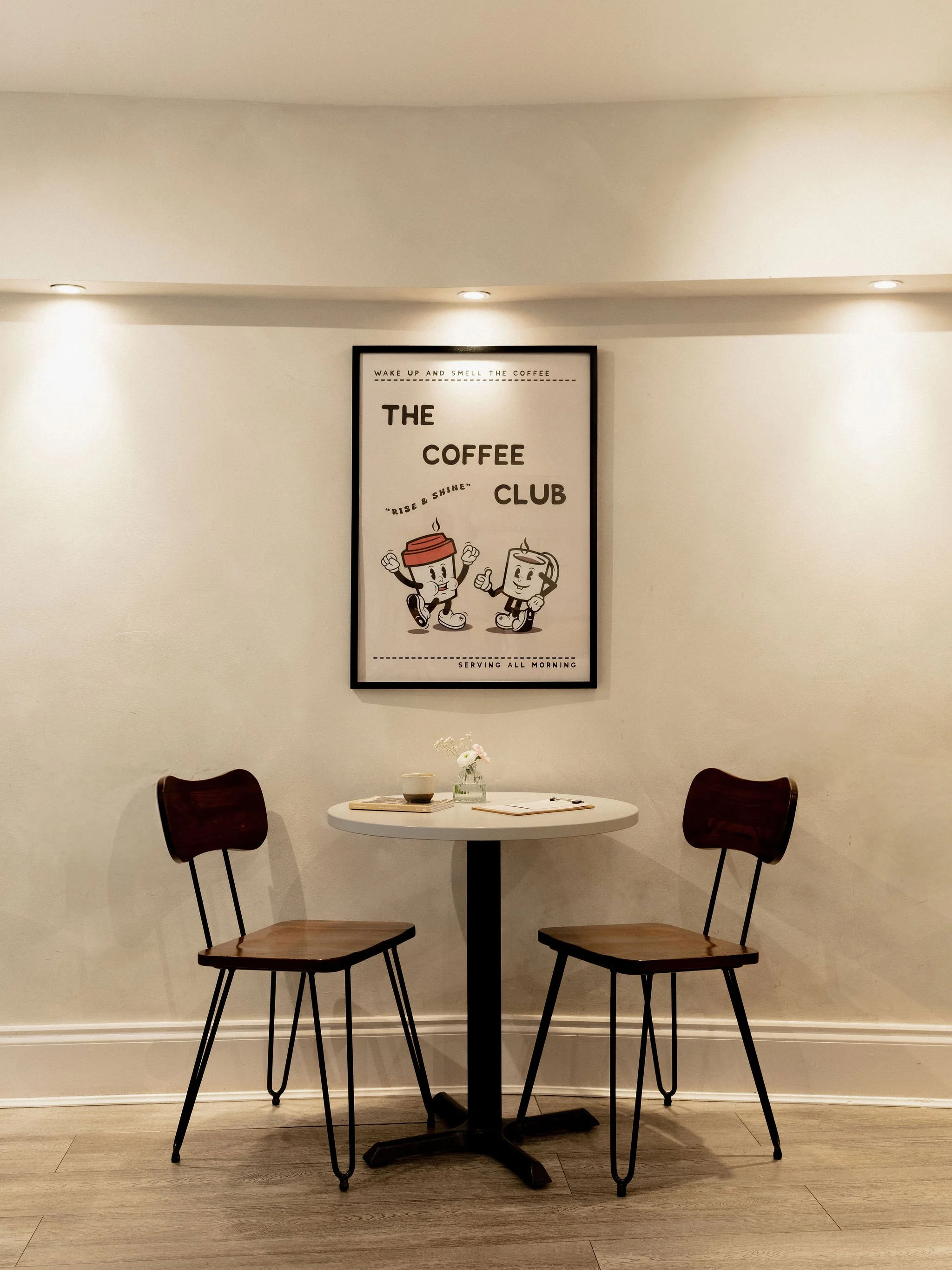 A small round table with a white top and black base, accompanied by two wooden chairs with black metal legs, set against a white wall. A framed poster featuring cartoon coffee cups and text about a coffee club hangs on the wall. The table has a small flower vase, a cup, and a closed notebook.