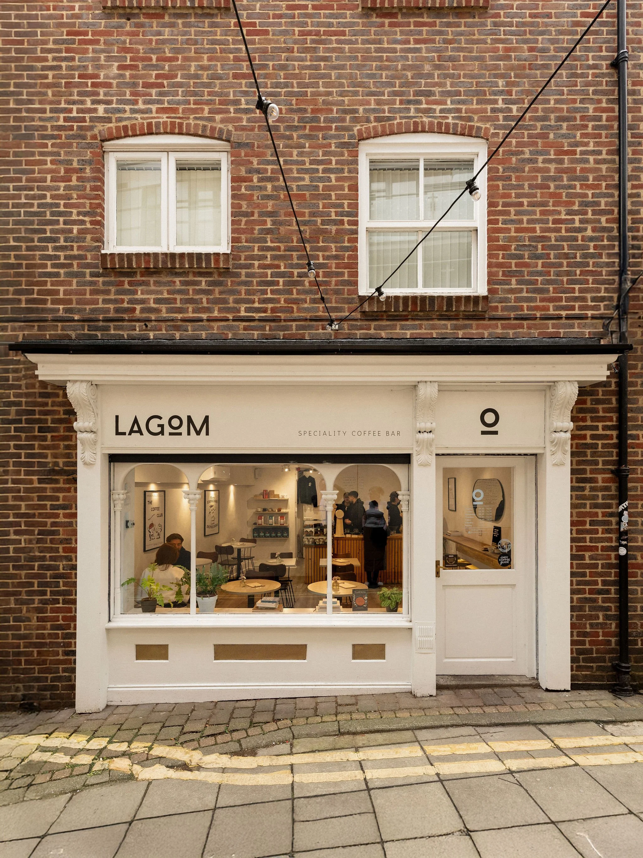 Exterior view of a coffee shop called LAGOM with large front window, small shop door, and brick building. Inside, people sit at tables and baristas prepare drinks.