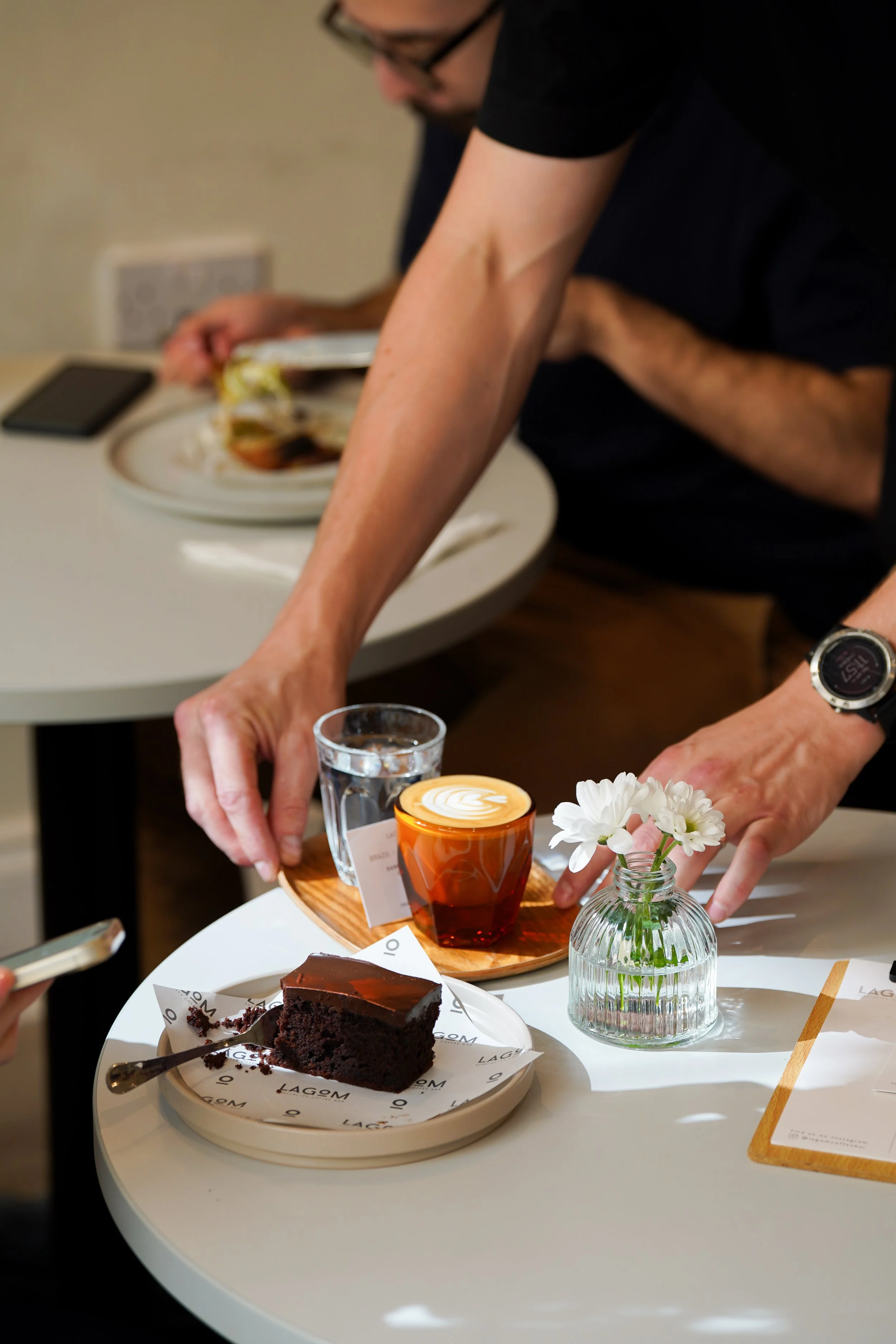 A person's hands reach for a slice of chocolate cake on a white table, with a latte in a red cup, a glass of water, and a small vase with white flowers nearby.