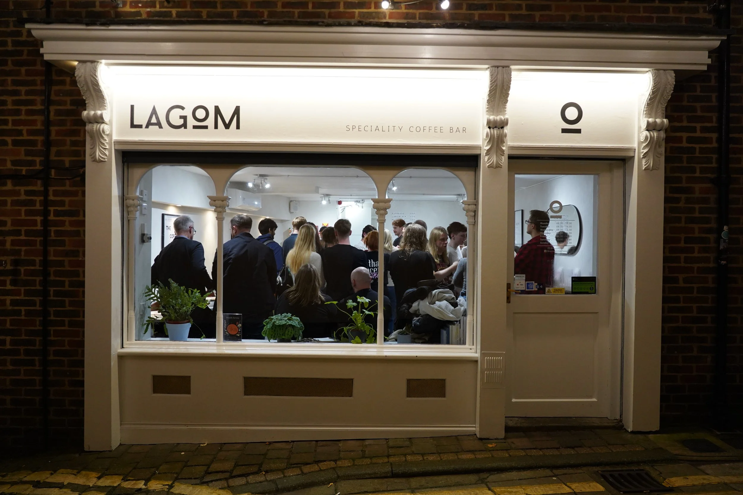 Exterior of a coffee shop named 'LAGOM' with large front window showing customers inside and a door to the right. The shop is lit up at night, with brick wall on the sides and decorative white trim around the window and door. Inside, customers are gathered, some standing in line.