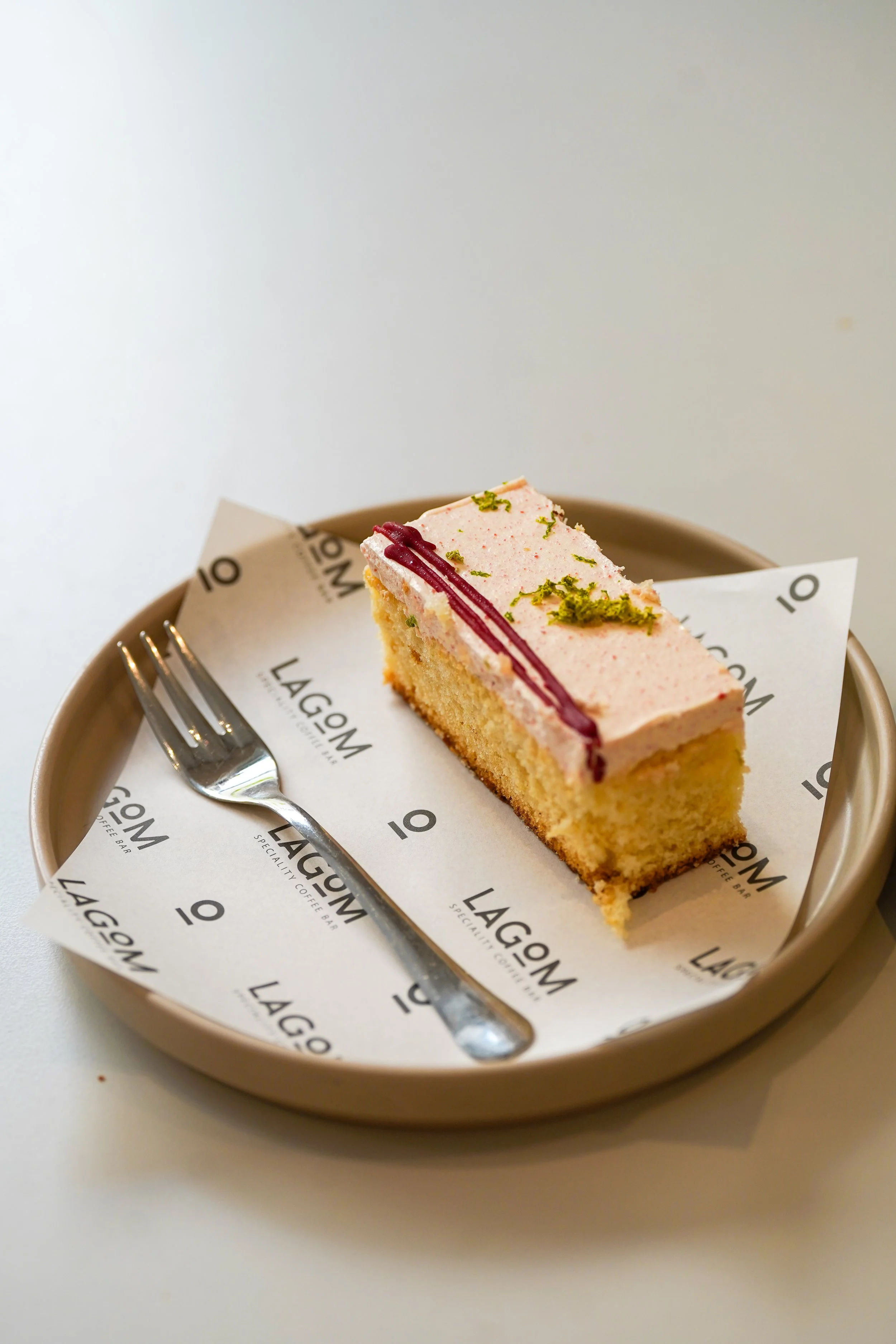 A slice of pink frosted cake with red and green garnishes on a beige plate, accompanied by a silver fork, on branded paper from Lagom coffee bar.