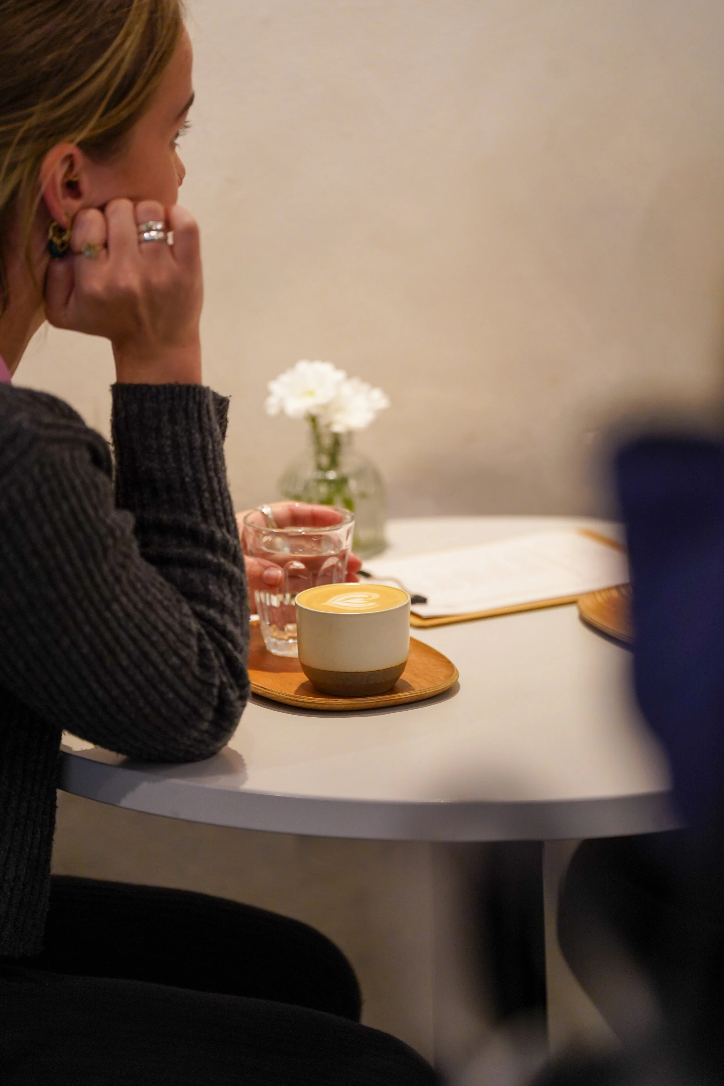 A woman with blonde hair sits at a white table in a cafe, resting her head on her hand. On the table, there is a cup of coffee with latte art, a glass of water, and a small vase with white flowers.