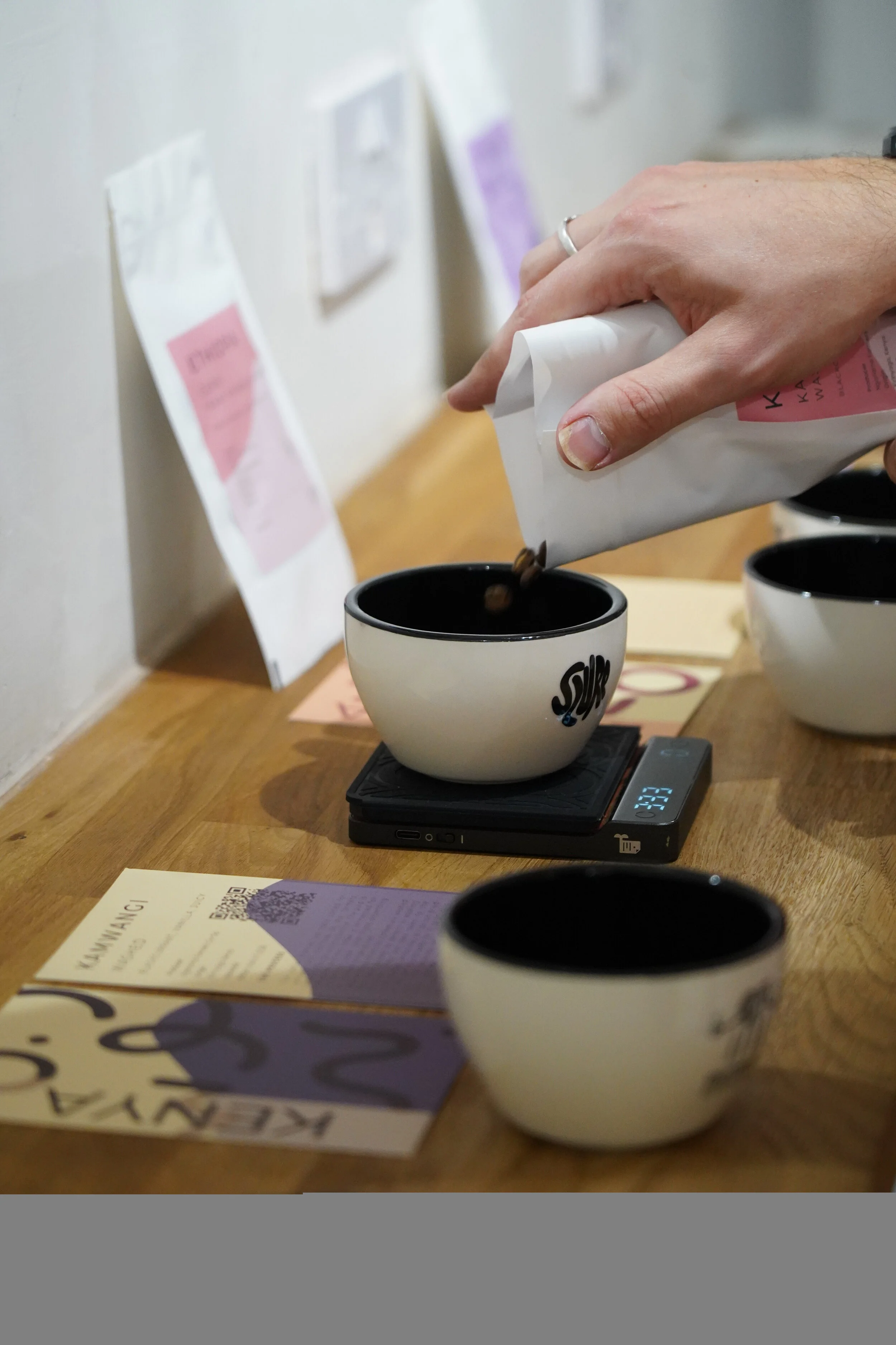 A person is pouring coffee into a black and white ceramic cup on a wooden counter, with other cups, a scale, and informational pamphlets nearby in a coffee shop setting.