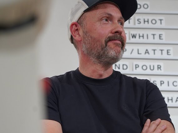 A man with a beard and mustache wearing a black t-shirt and a baseball cap, standing in front of a menu board with white letter tiles.