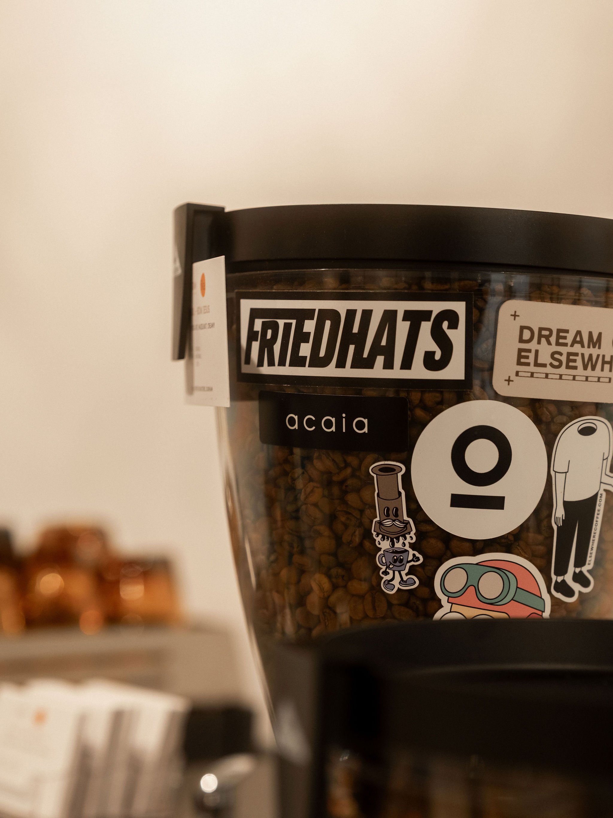 Close-up of a container of coffee beans with various colourful stickers, including one with a circular Lagom logo and a cartoon character, on a countertop with blurred background.