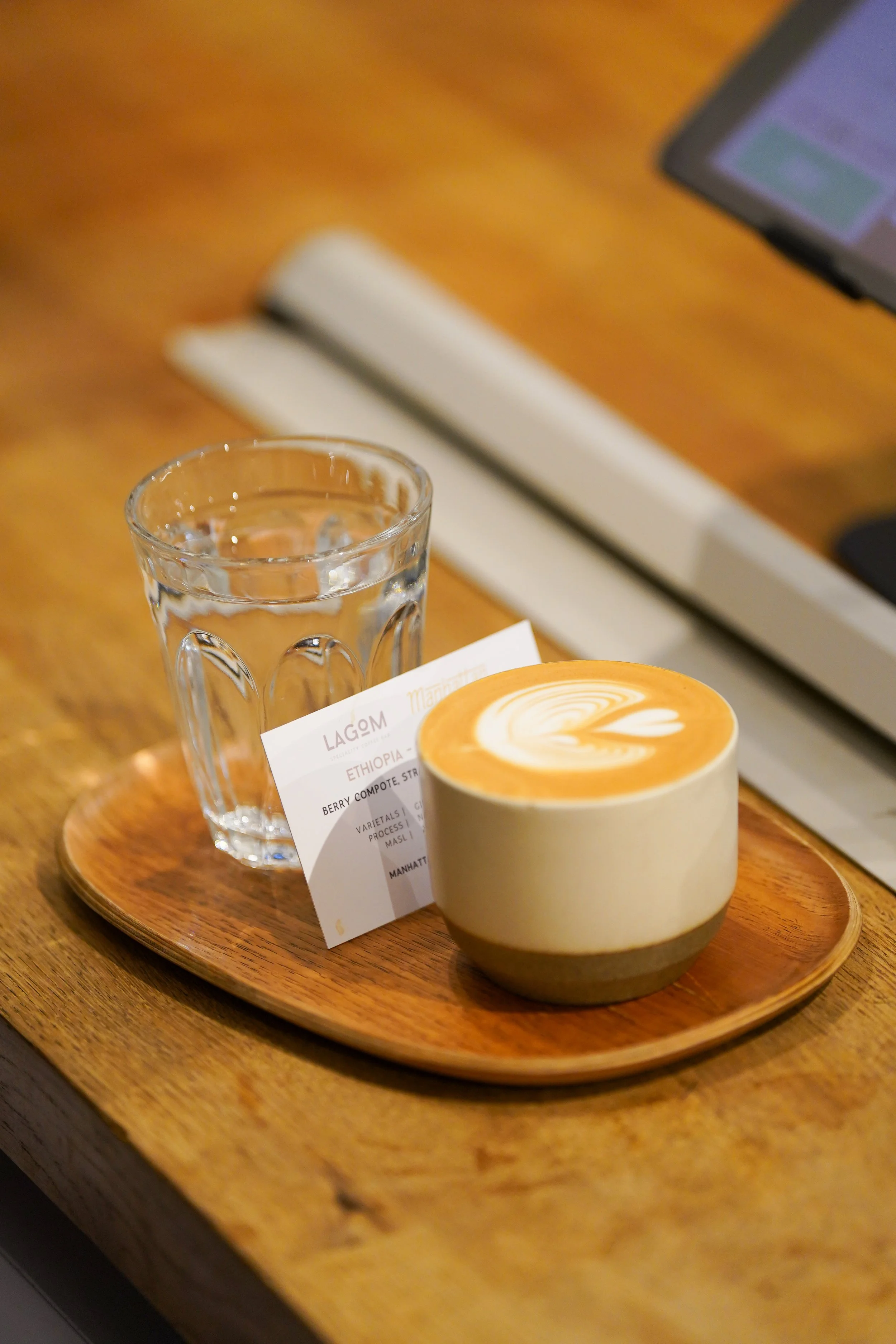 A wooden tray with a glass of water, a cup of latte with latte art, and a printed order slip, on a wooden table in a coffee shop.