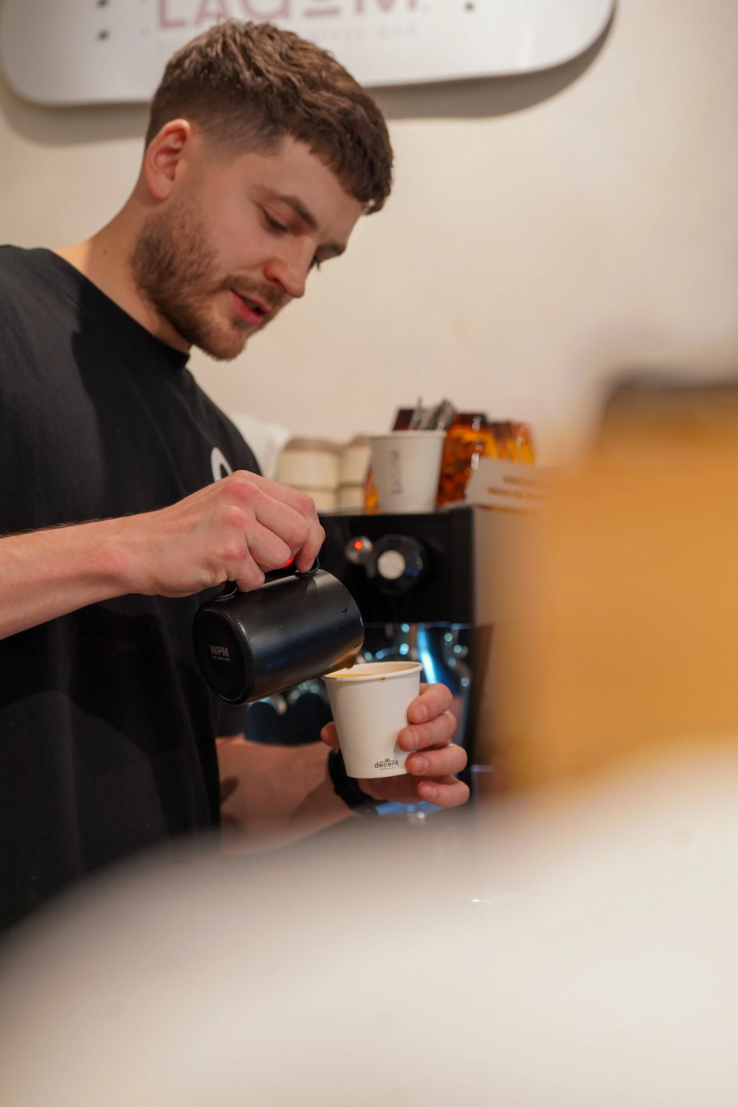A barista pours coffee from a black pitcher into a white disposable cup at a coffee shop. The barista has short brown hair, a beard, and wears a black shirt. Background includes coffee equipment and a blurred sign.