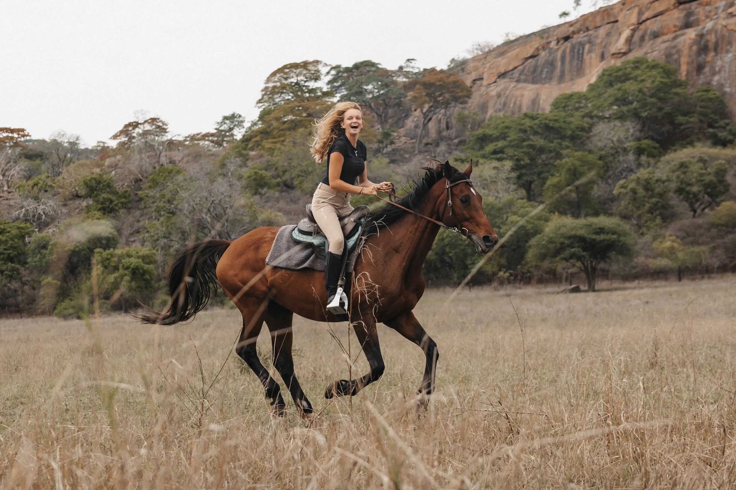 Volunteer riding a brown horse in a grassy field with trees and rocky hills in the background.