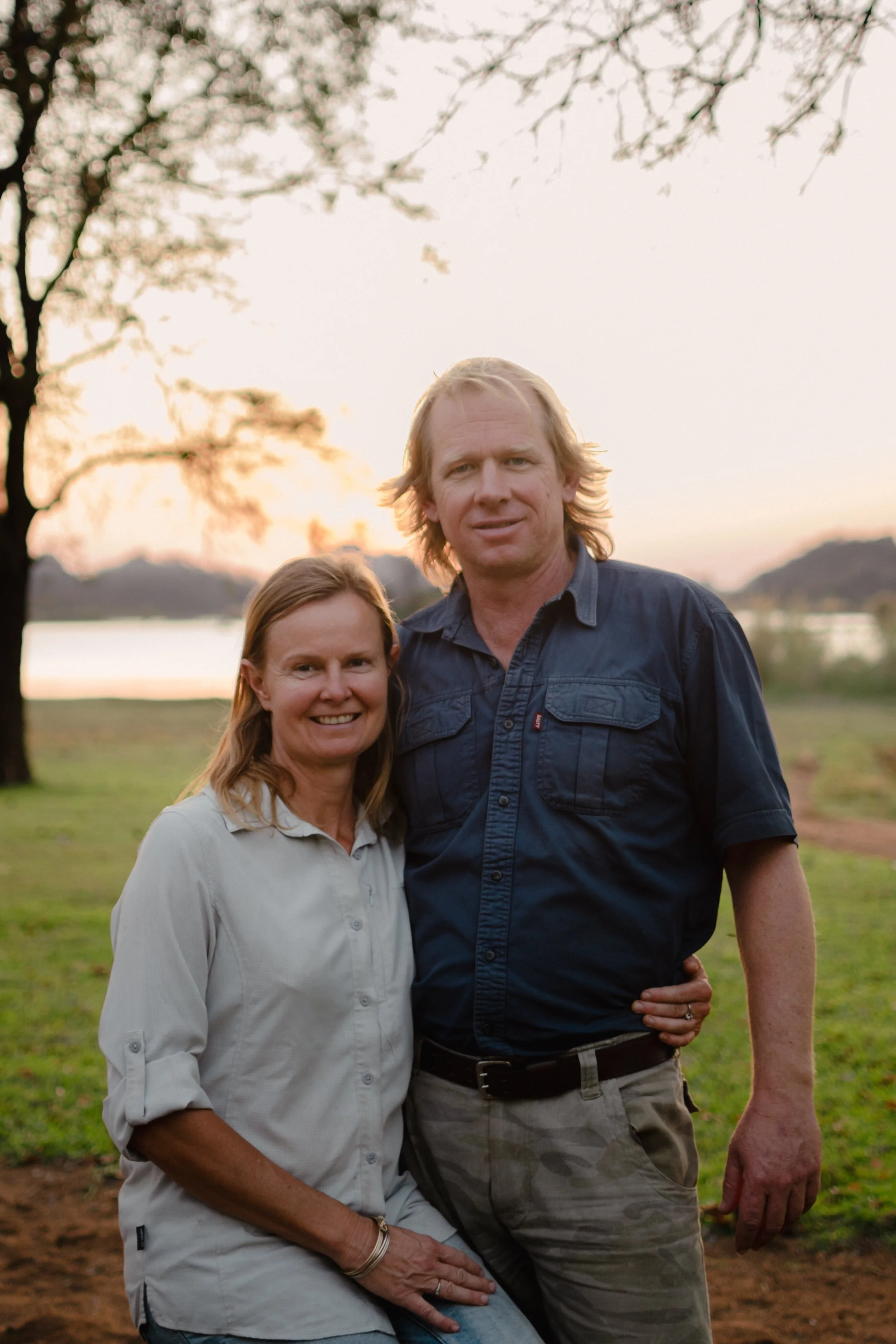 A couple standing outdoors near a lake during sunset with trees in the background.