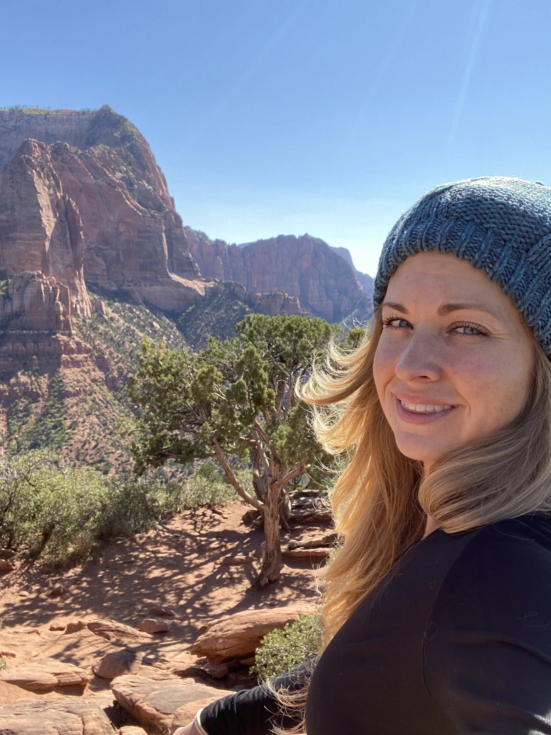 A woman with blonde hair wearing a blue knit beanie smiling outdoors in a desert landscape with red rock cliffs and sparse trees.