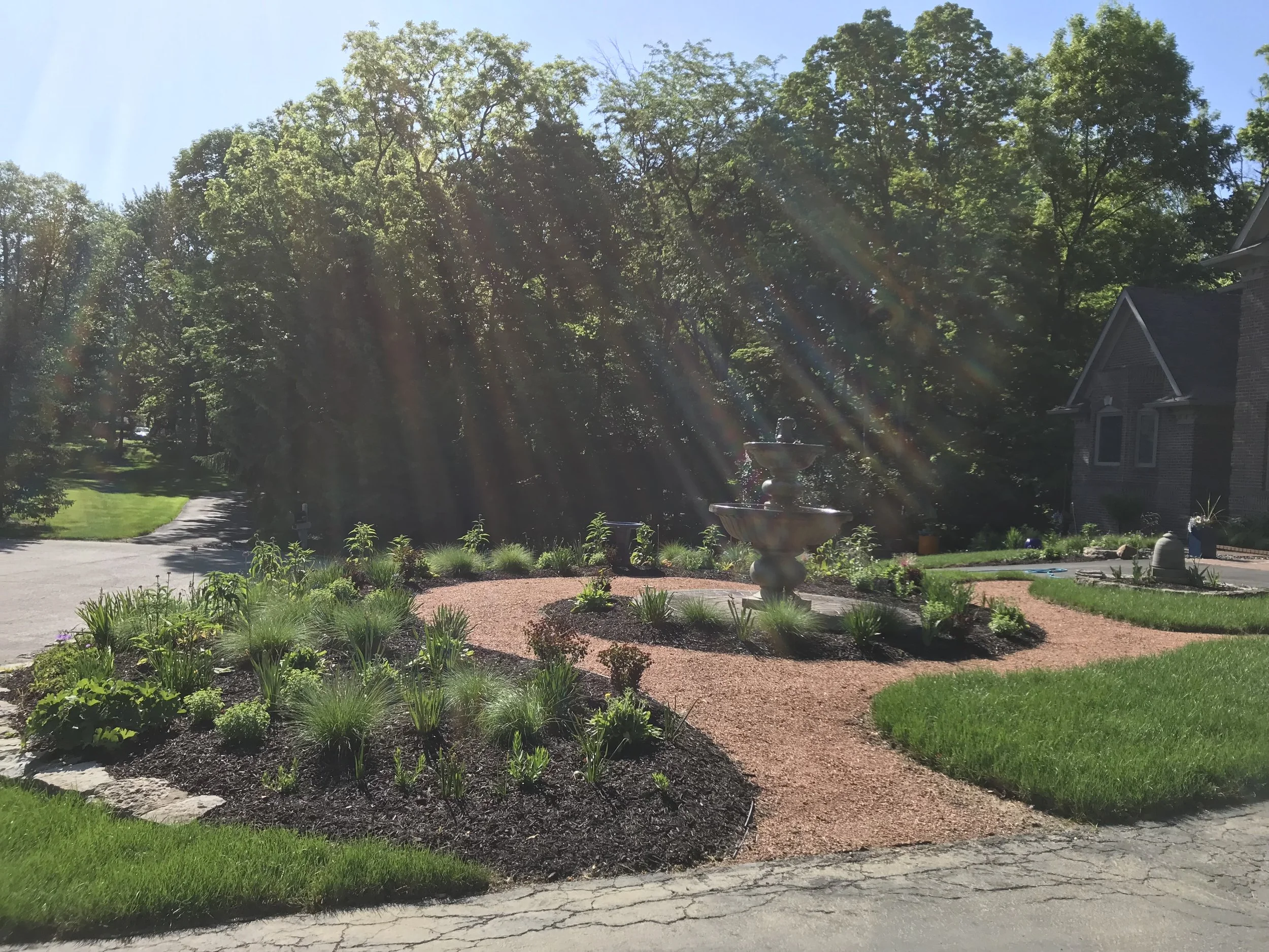 A landscaped front yard of a house with a multi-tiered fountain, green grass, mulch beds with plants, and trees in the background under a clear blue sky with sunlight rays.