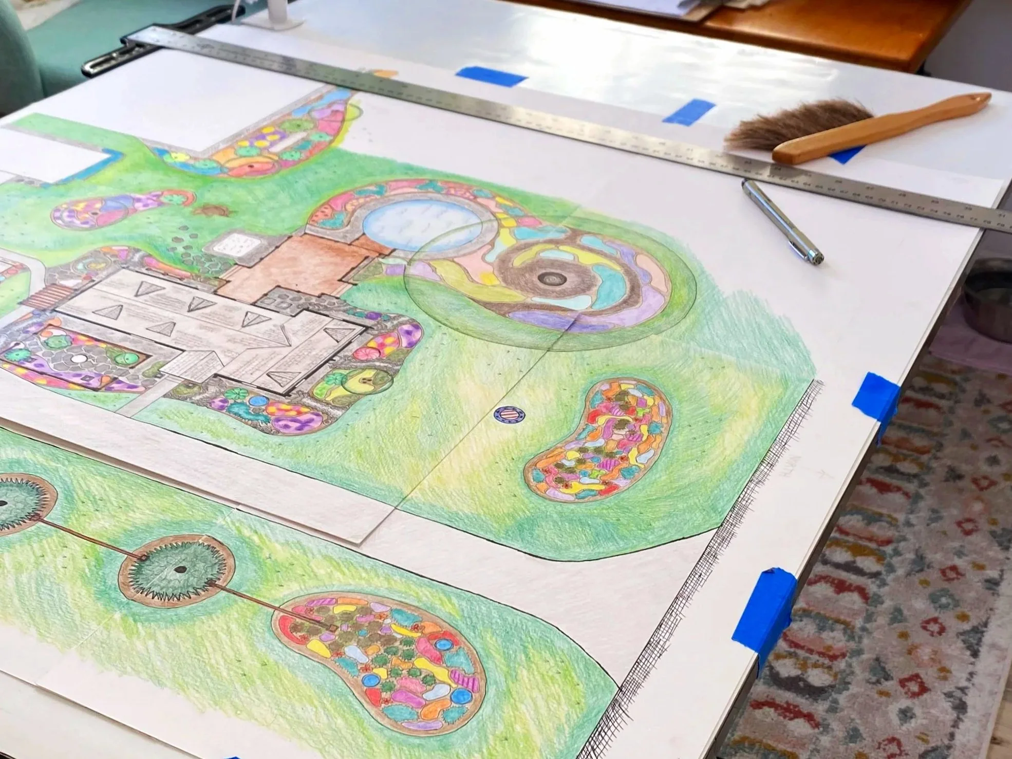 Colorful landscape design plan on a large sheet of paper, with various shapes, pathways, and landscaping elements, taped to a table with tools and a ruler.