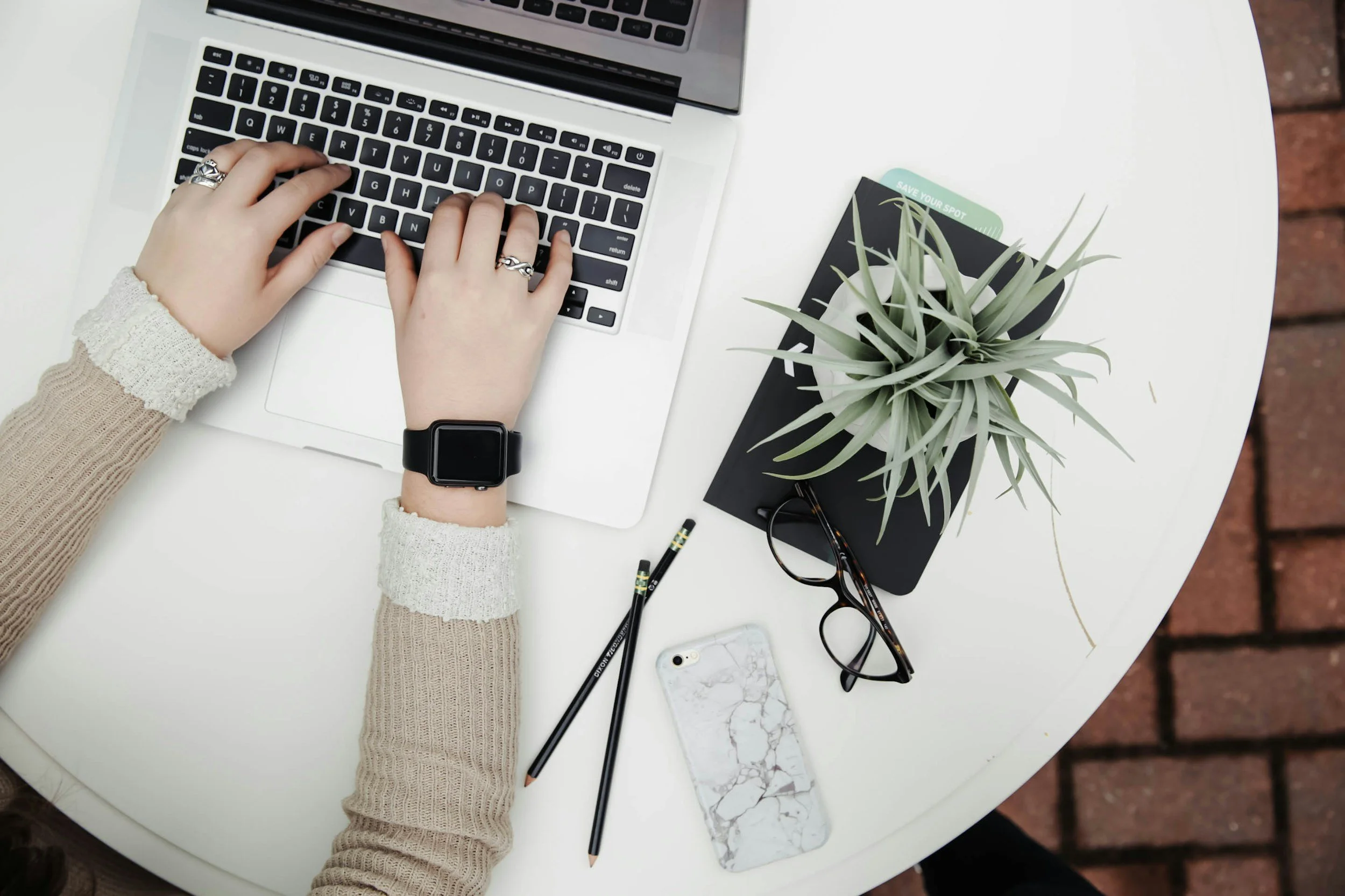 Person typing on a laptop at a round white table with a potted plant, smartphone, black glasses, and pencils.