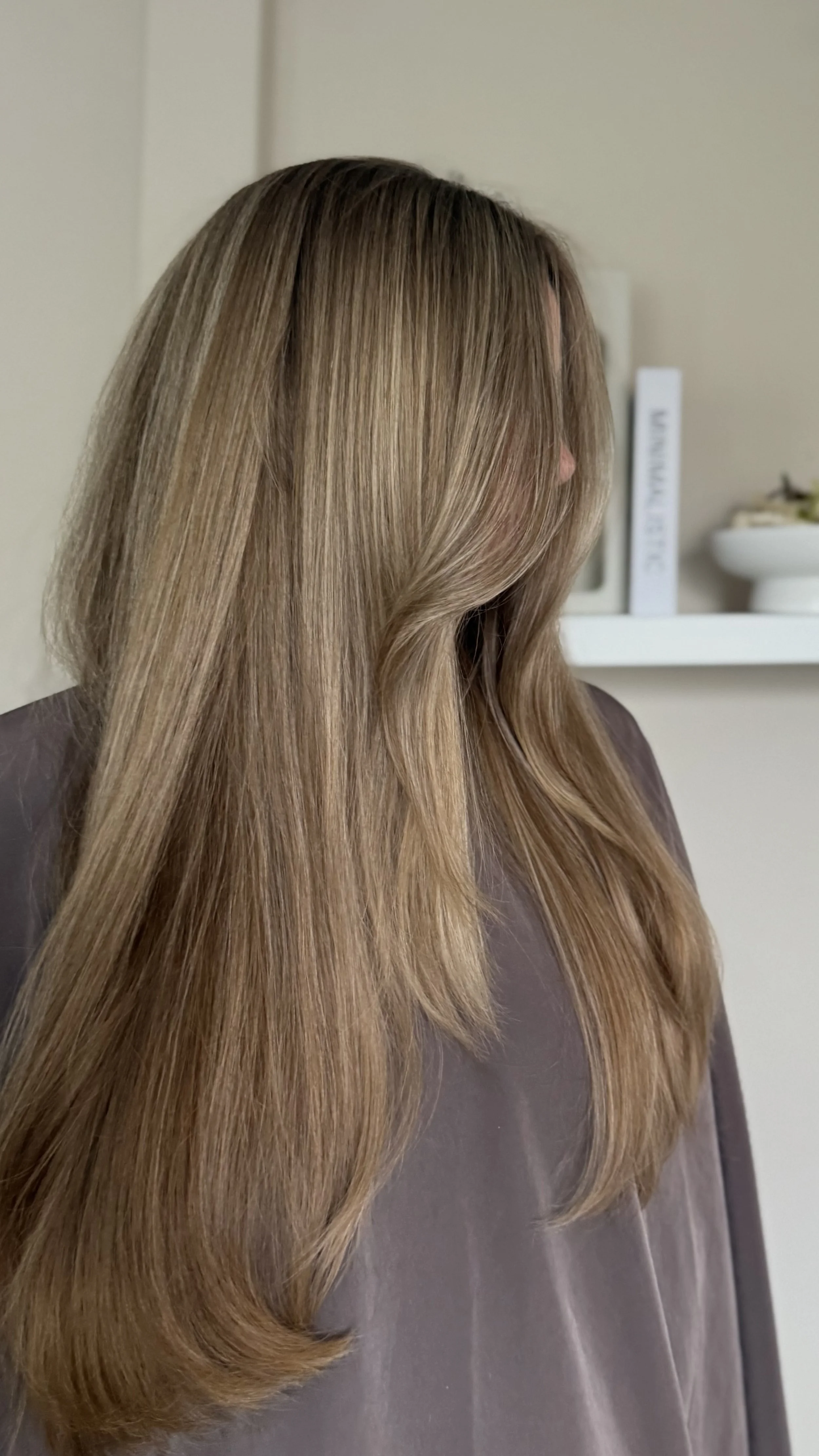 A woman with long, straight, light brown hair wearing a gray top, standing indoors against a beige wall with white shelves in the background.