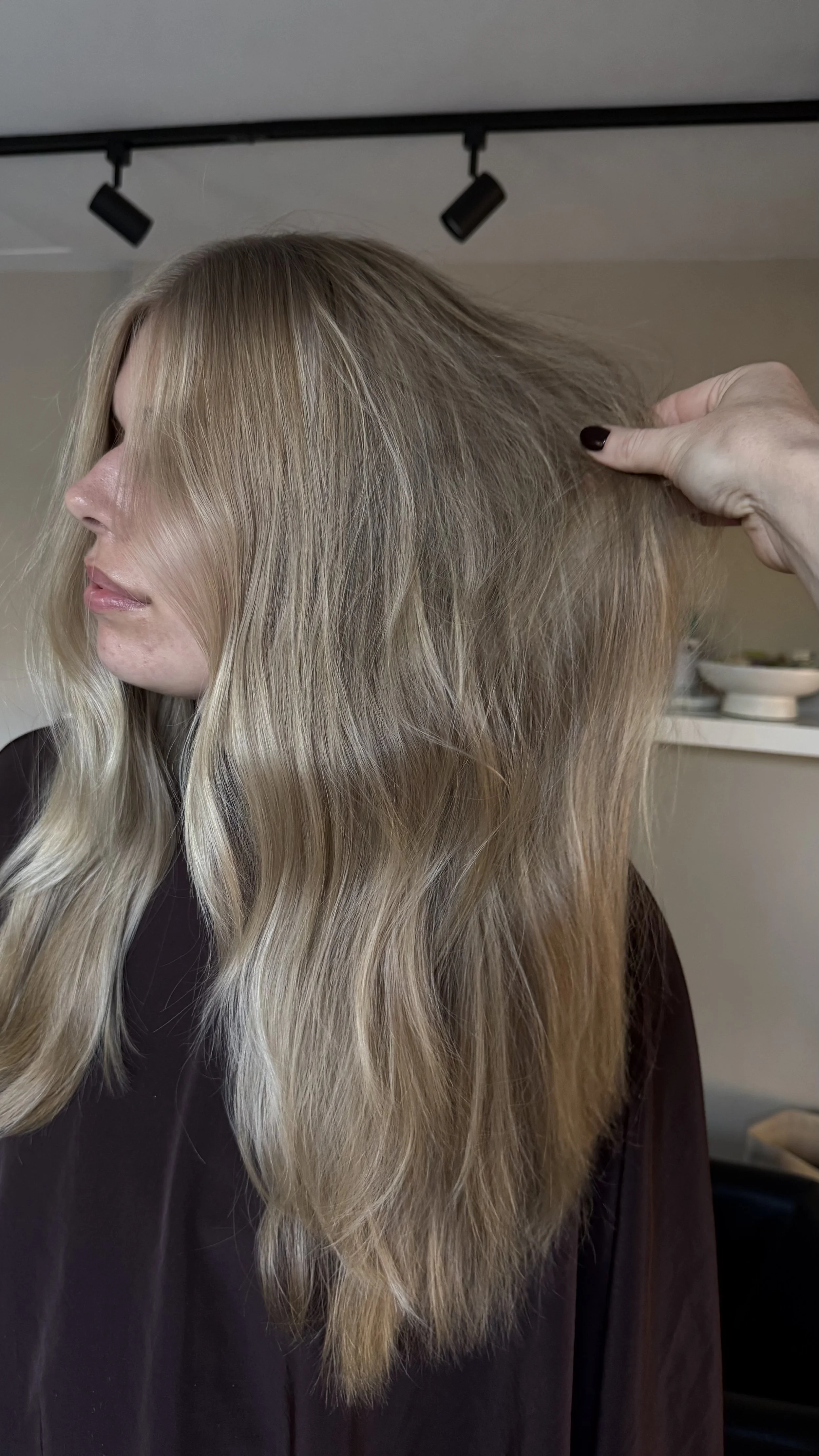 Close-up of a woman with layered, blonde, wavy hair in a salon. She is wearing a dark brown top, and the background shows salon shelves with various hair products.