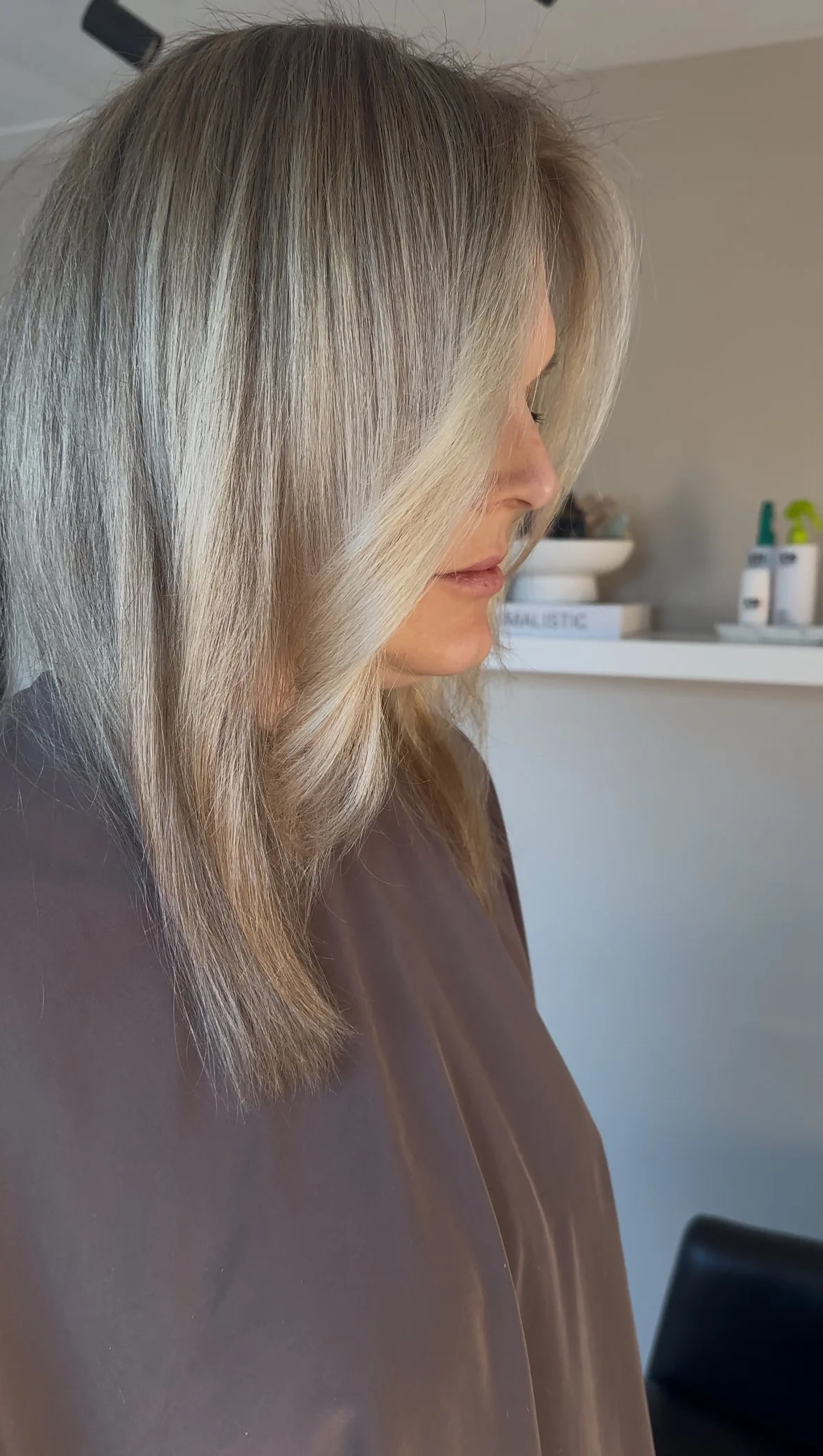 A woman with blonde, layered hair sits in a salon chair, looking down slightly, wearing a brown salon cape.