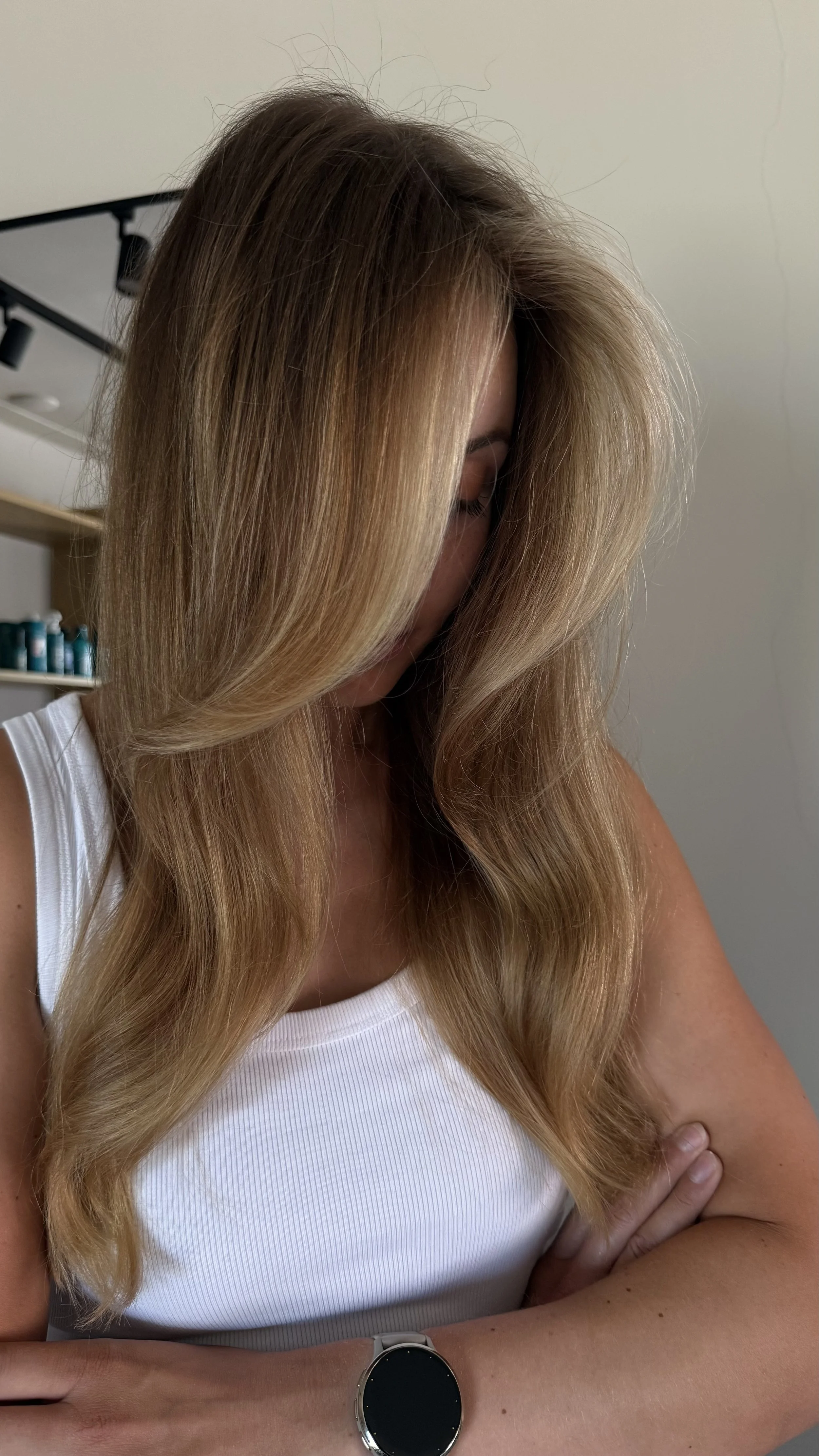 A woman with blonde, layered hair sits in a salon chair, looking down slightly, wearing a brown salon cape.