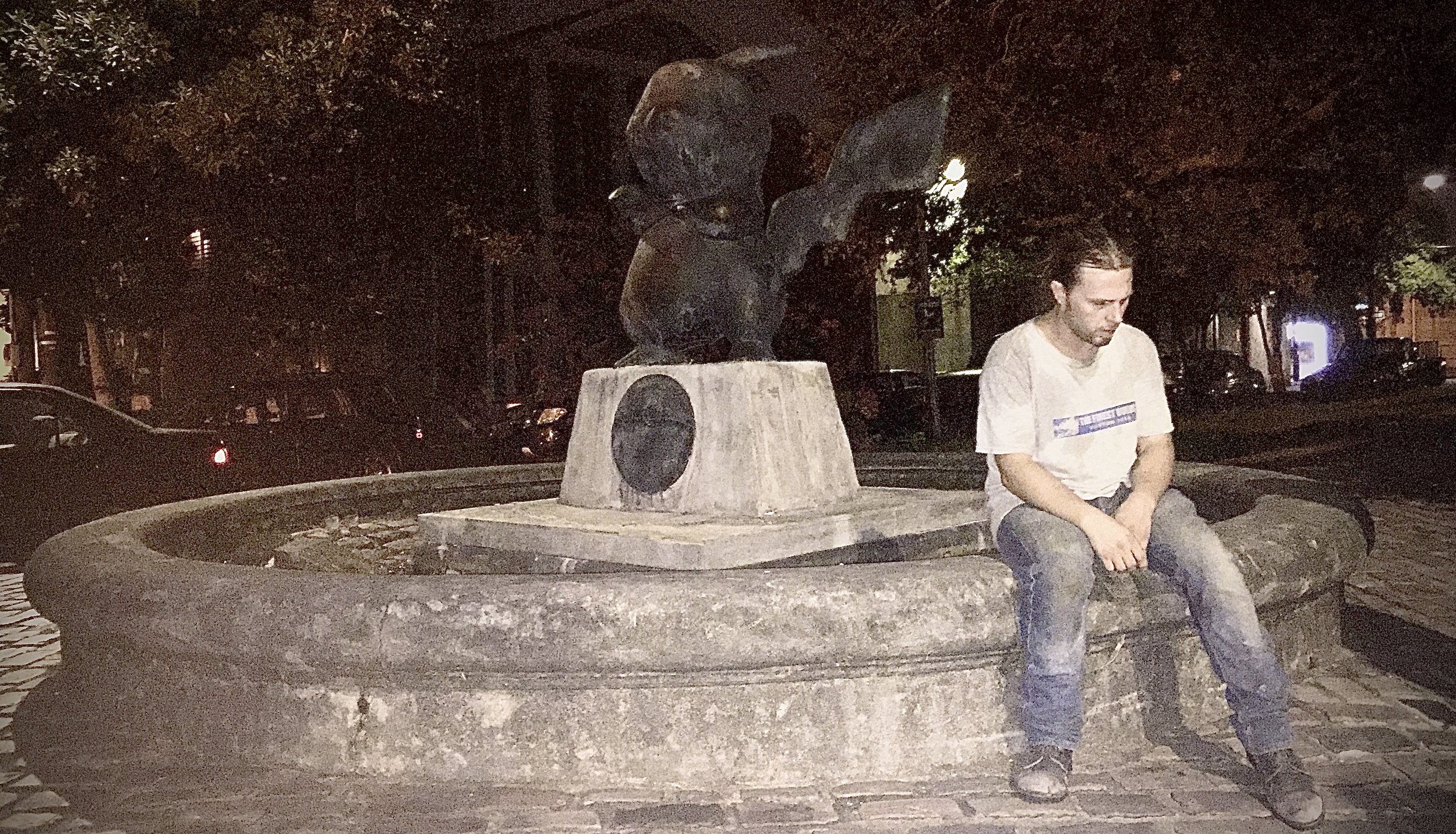 A man sitting alone on a stone fountain in a park at night, with a statue of a rabbit in the background and parked cars in the distance.