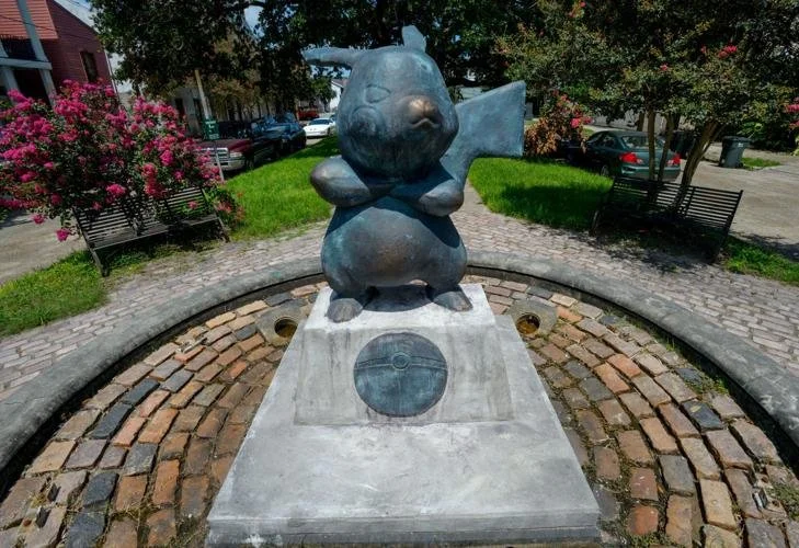 Bronze statue of a fish with a smiling face, mounted on a square concrete pedestal in a park with benches, trees, and pink flowers.