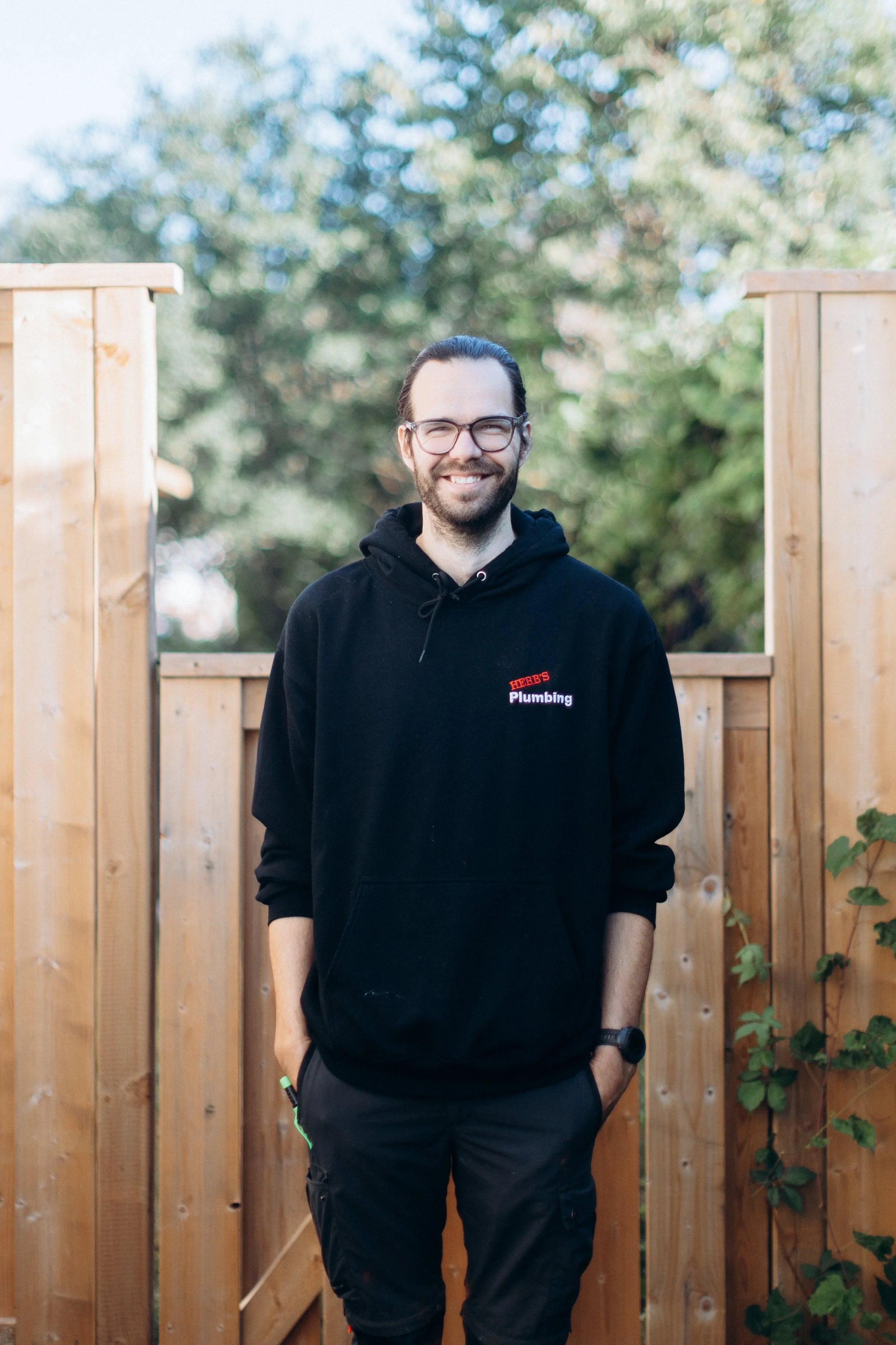 A man smiling in black hoodie smiling, wearing glasses and long hair tied back. standing in front of a wooden fence and greenery in the background