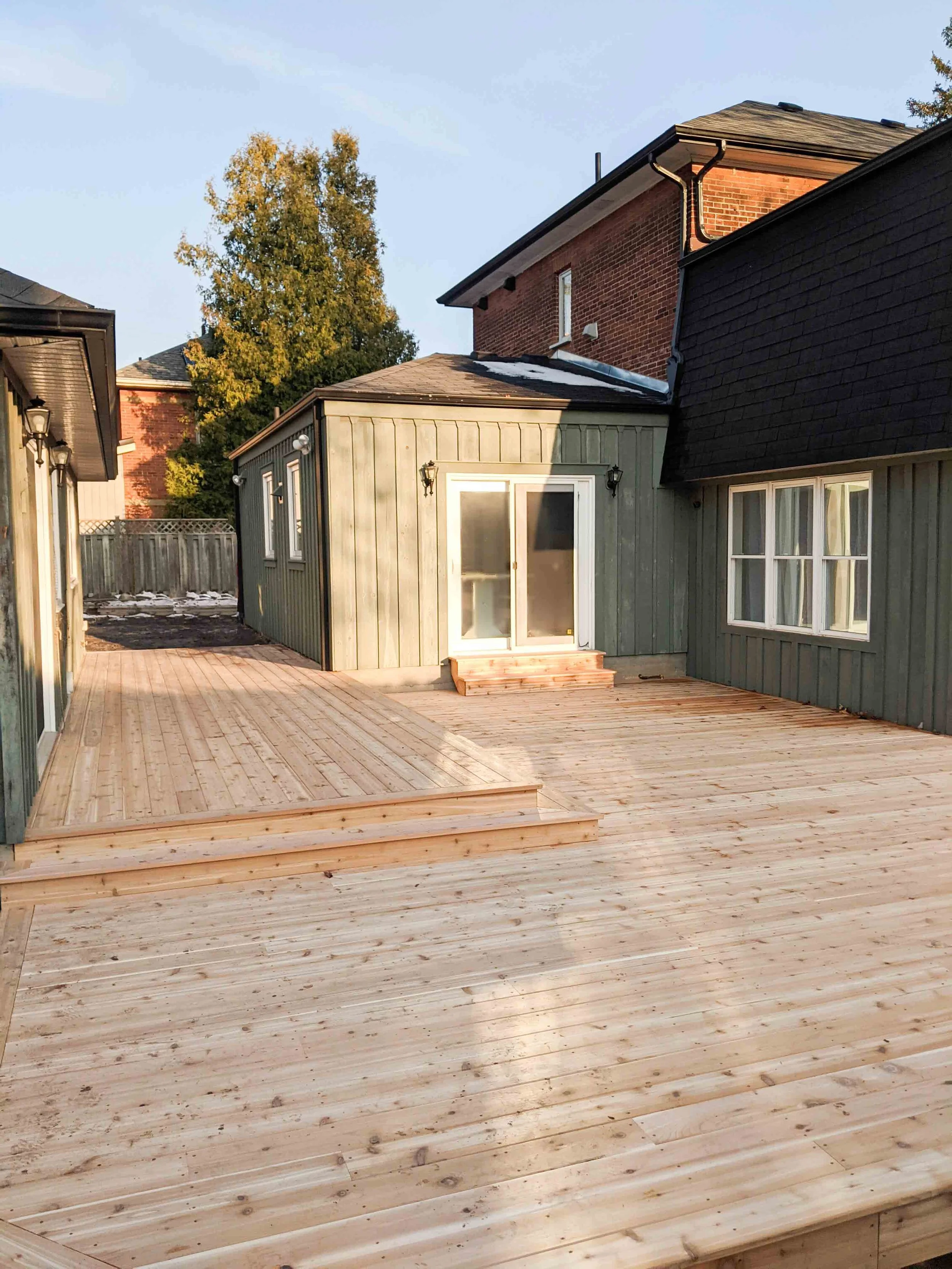 Newly built wooden deck outside a house with sliding glass doors and green exterior siding, in a residential backyard with neighboring houses and trees in the background.