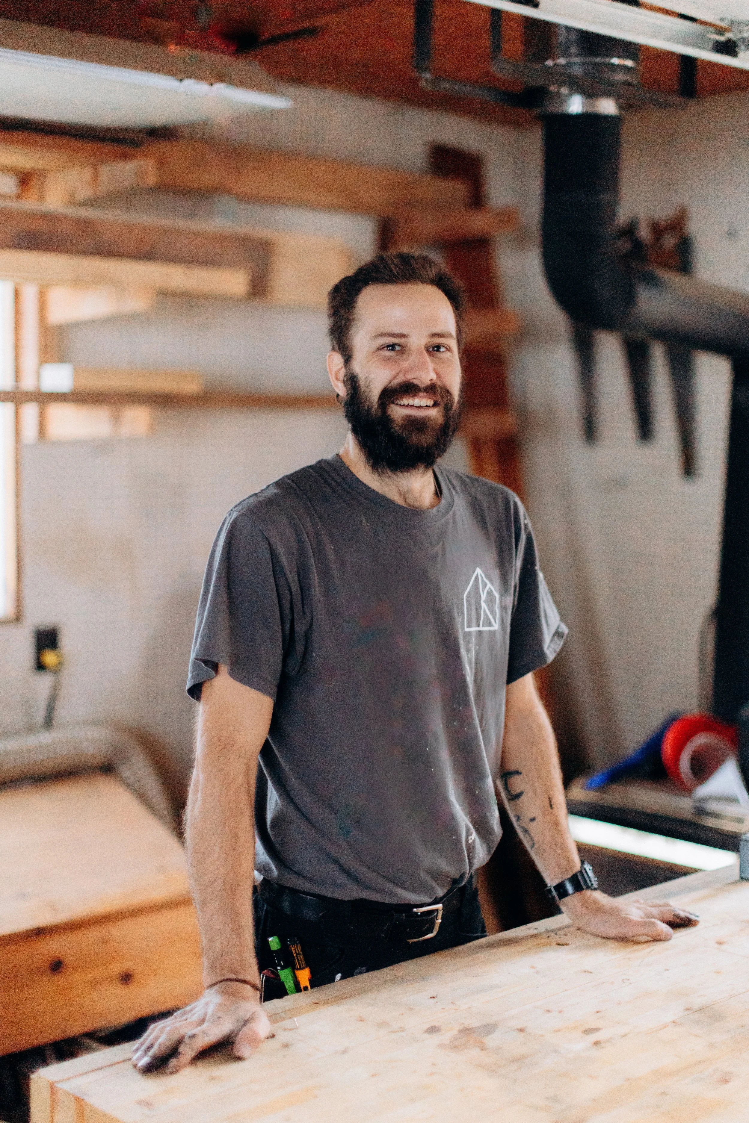 Close-up of a man with dark curly hair, glasses, a beard, and a nose ring, wearing a dark green sweater, looking slightly to the side with a thoughtful expression.