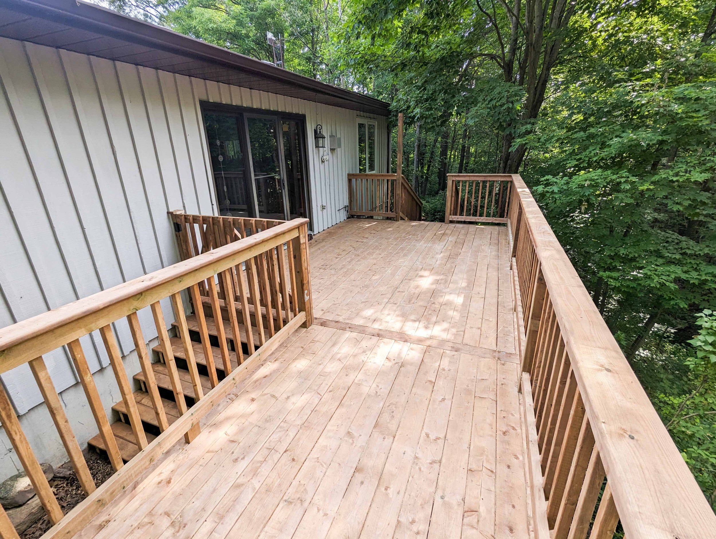 Photo of a new wooden deck attached to a house, overlooking a wooded area, with stairs leading down from the deck.