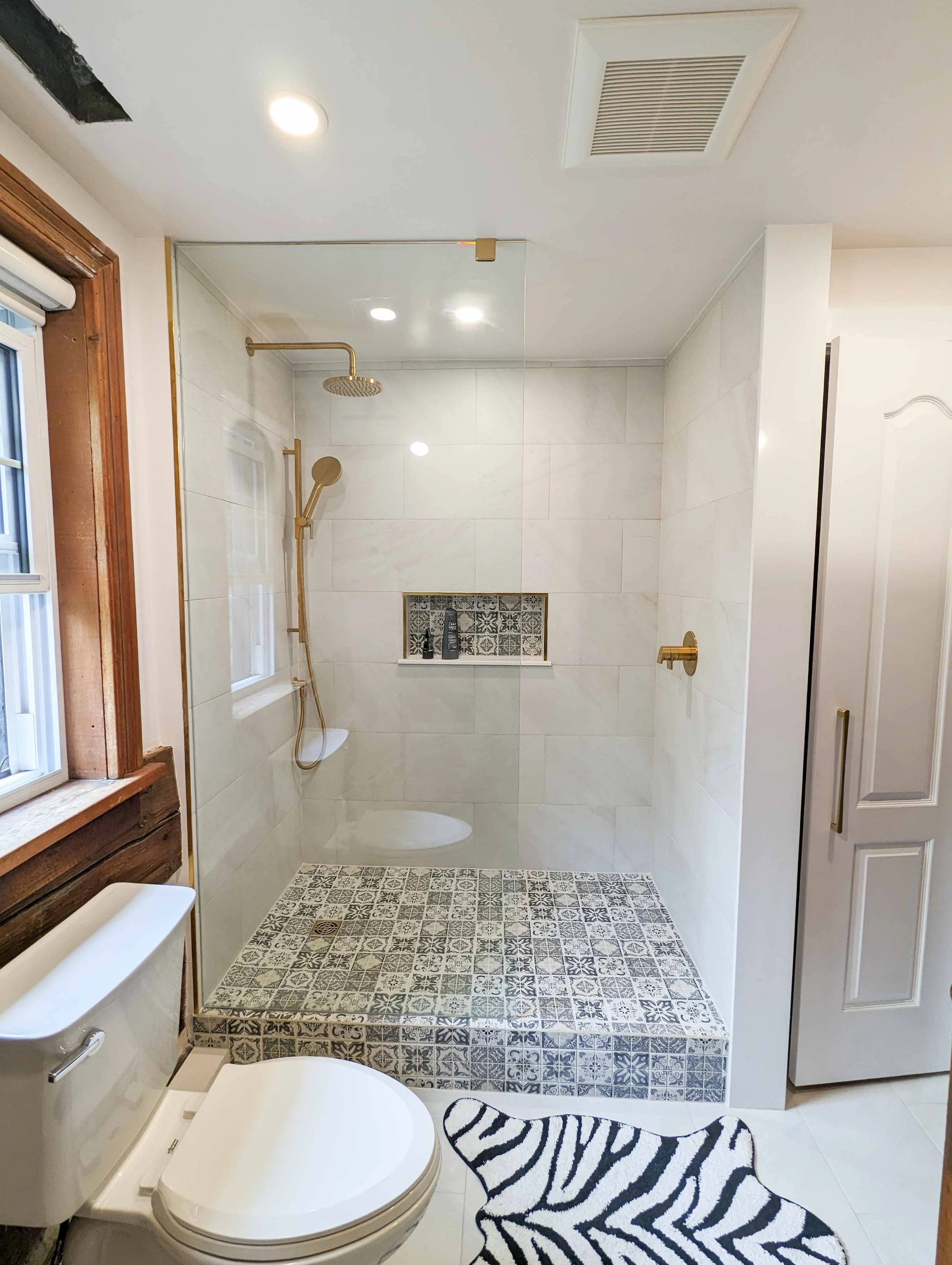 Modern bathroom with a walk-in shower featuring a glass door, gold fixtures, and patterned tile on the shower floor and back wall niche. To the left, there is a window, a toilet, and a zebra-print rug on the floor.