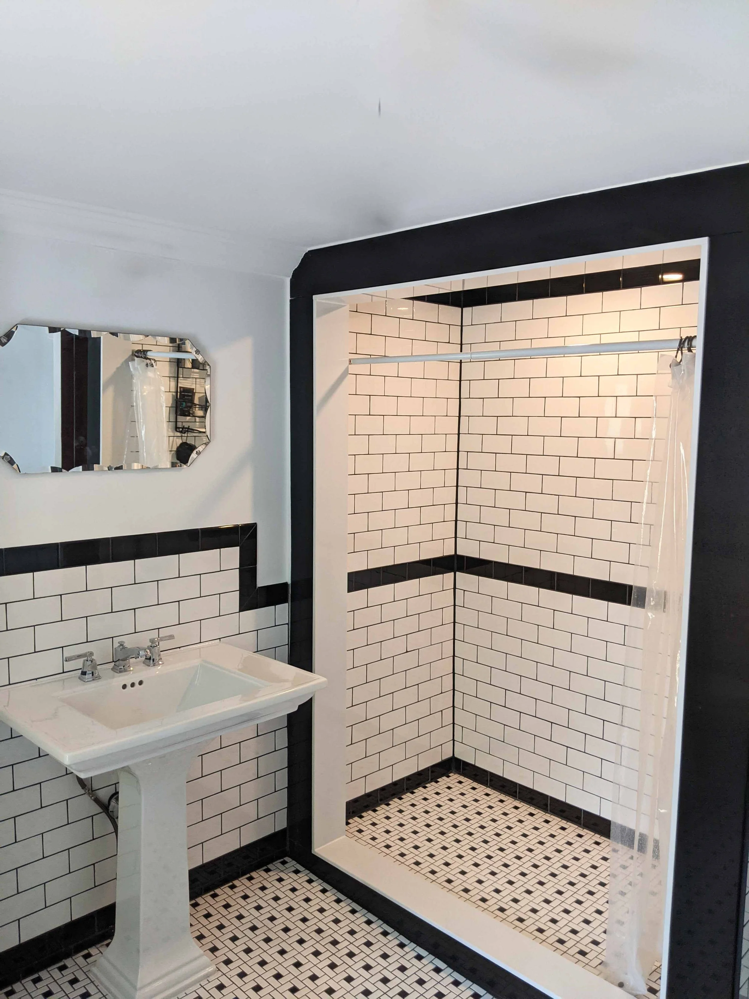 Bathroom with black and white tile design, including a corner shower with white subway tiles and black accents, a pedestal sink with a mosaic mirror, and a section of black wall around the shower area.