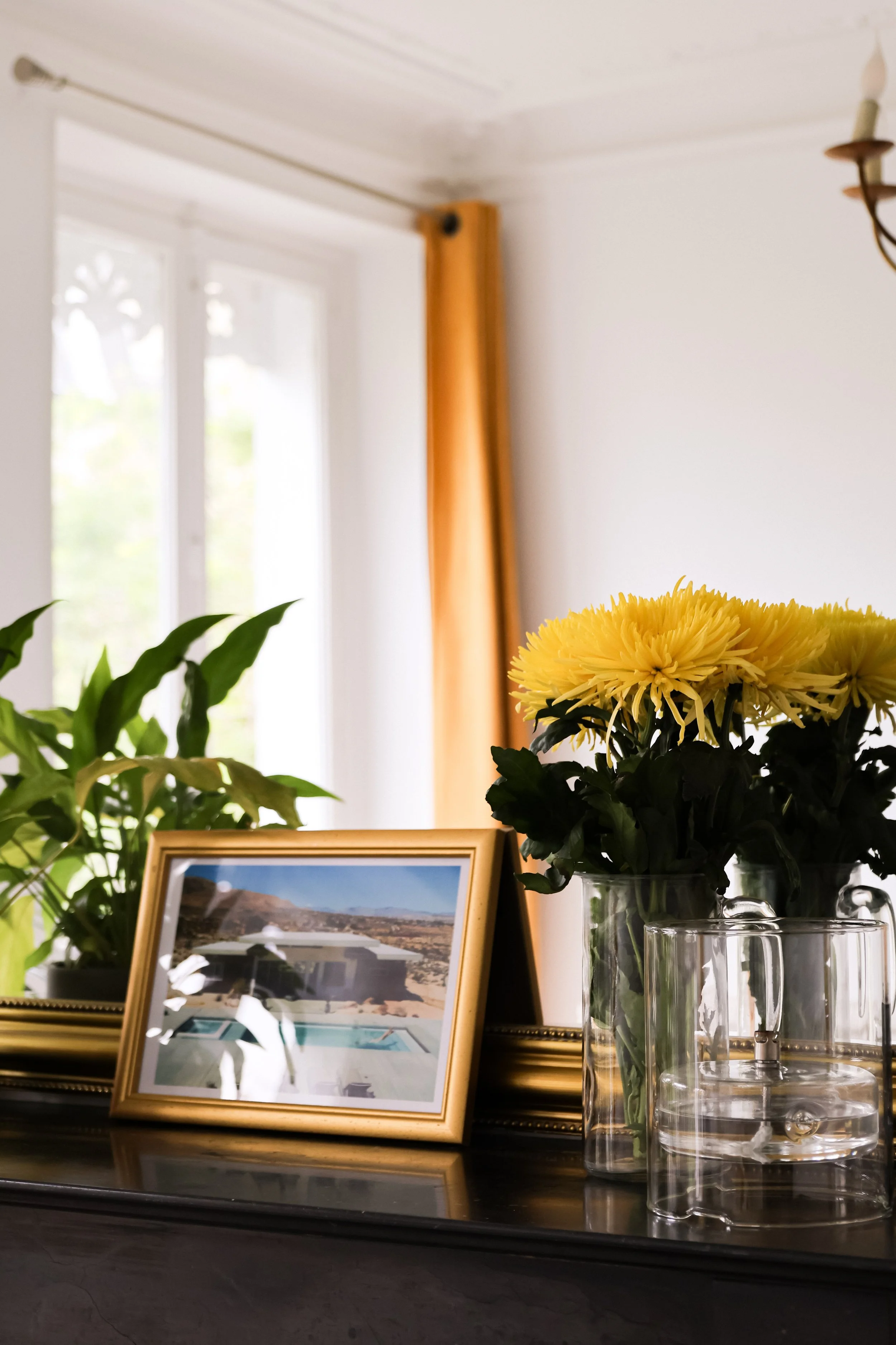 Vase avec des fleurs jaunes sur une surface en bois, photo encadrée, plante verte, fenêtre avec rideau orange en arrière-plan, intérieur lumineux.