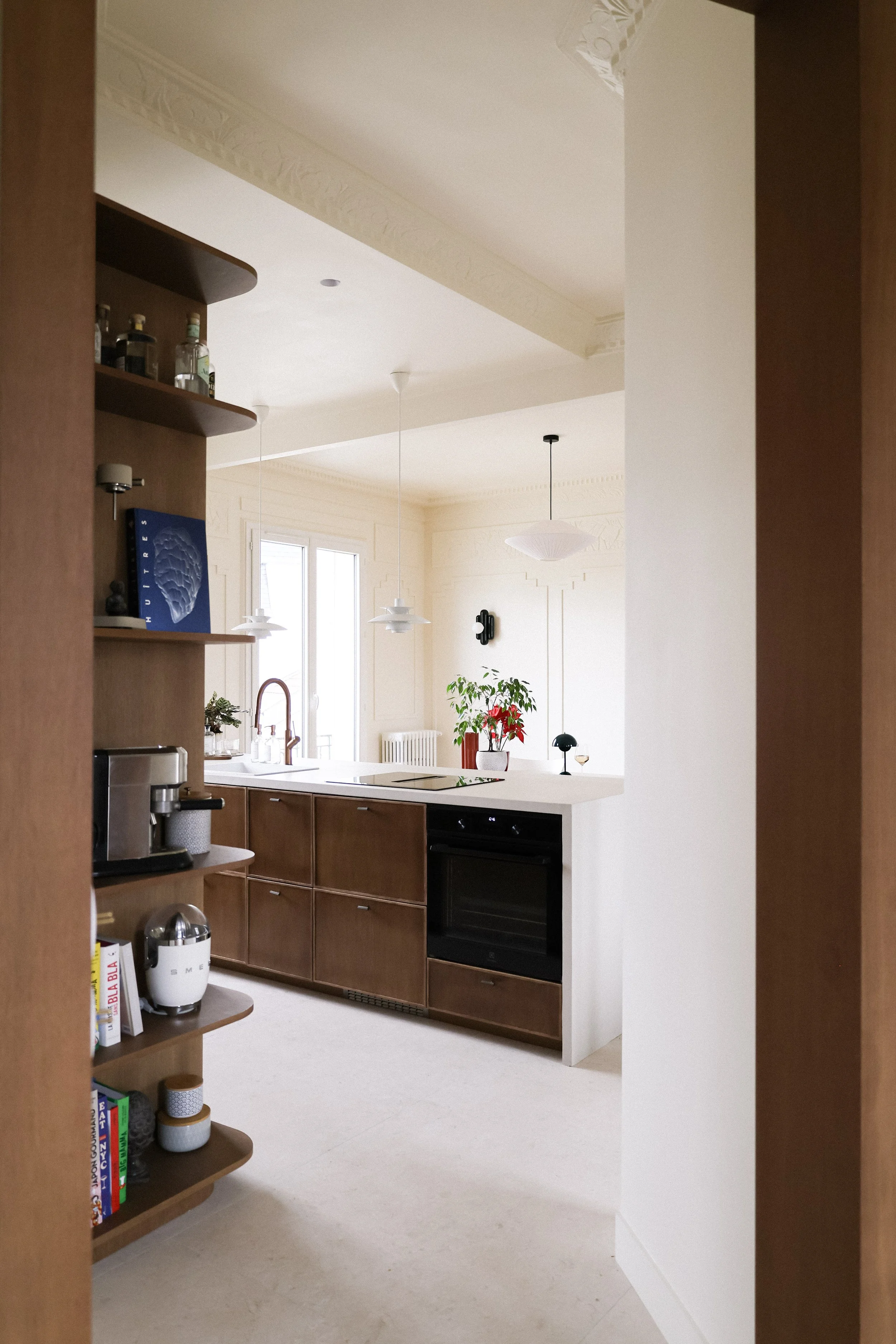 Une cuisine lumineuse avec des armoires en bois, un plan de travail blanc, une étagère avec des bouteilles d'alcool et des livres, une plante verte avec des décorations de Noël, et de grandes fenêtres laissant entrer la lumière naturelle.