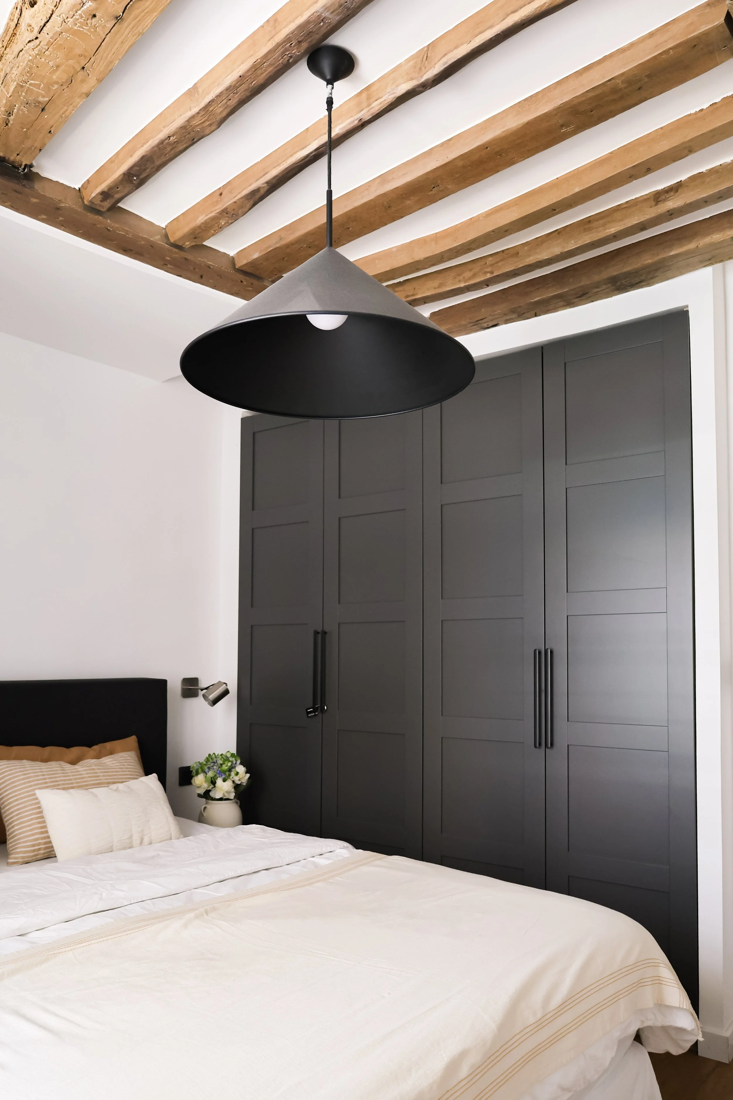 Chambre moderne avec un lit à draps blancs, oreillers beige et une grande armoire grise, plafonnier noir avec abajour, plafond avec poutres en bois apparentes.