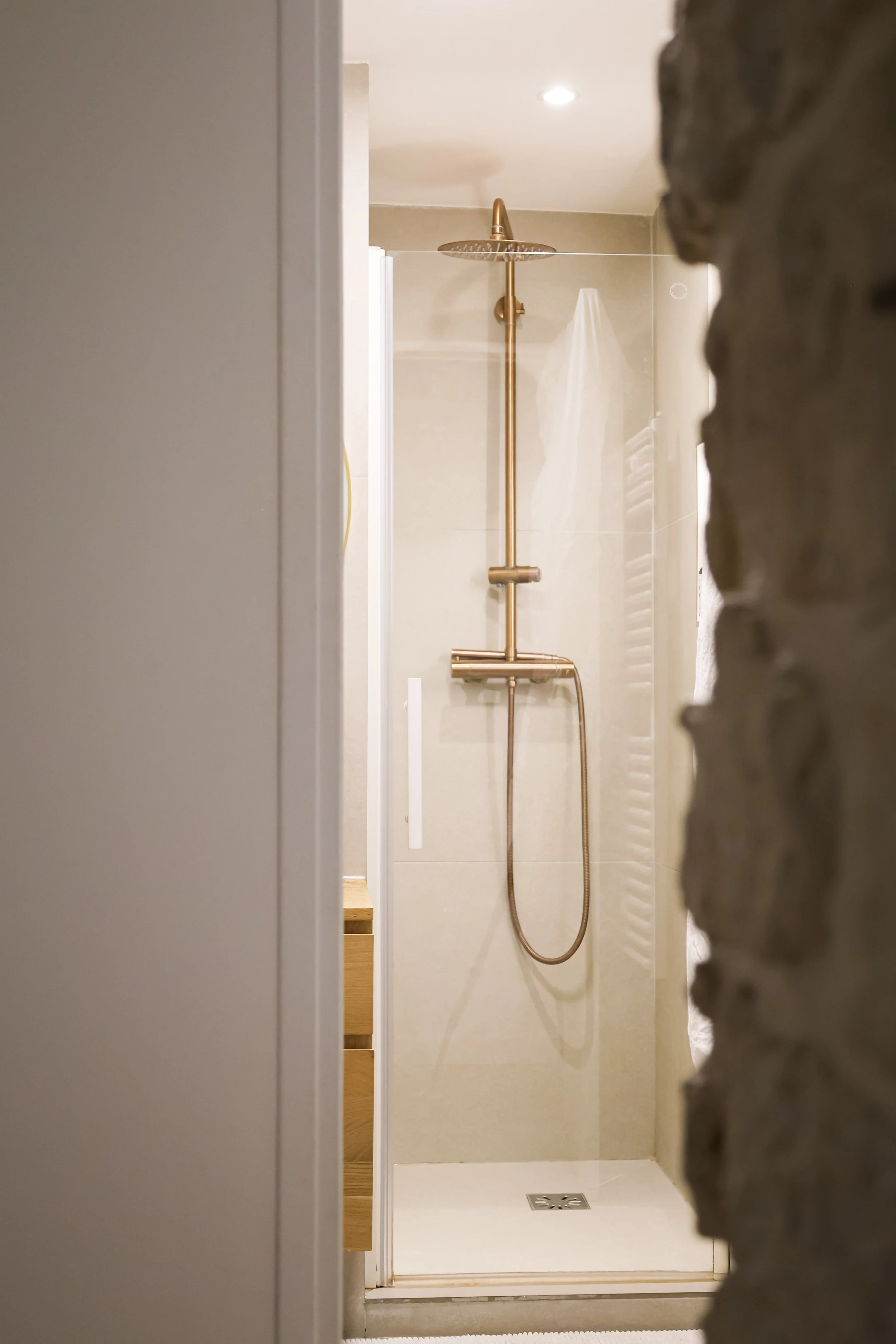 Douche à l'italienne avec pomme de douche en laiton, murs clairs et sol en carrelage beige, vue à travers une porte entrouverte, mur en pierre à droite.