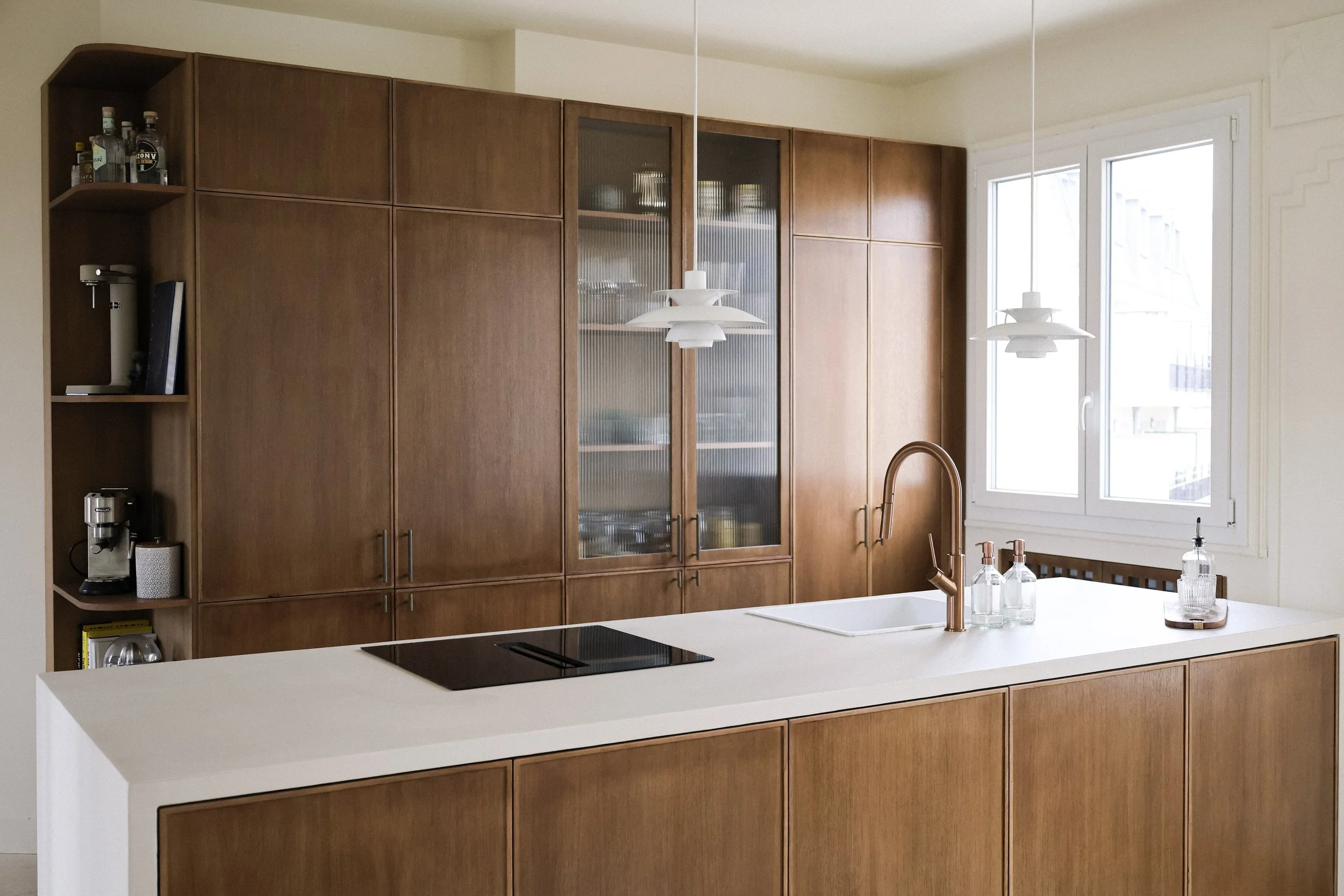 Une cuisine moderne avec un îlot central en bois et un plan de travail blanc, une fenêtre laissant entrer de la lumière naturelle, deux suspensions blanches au plafond, un évier en cuivre, et des bouteilles d'eau sur l'îlot.