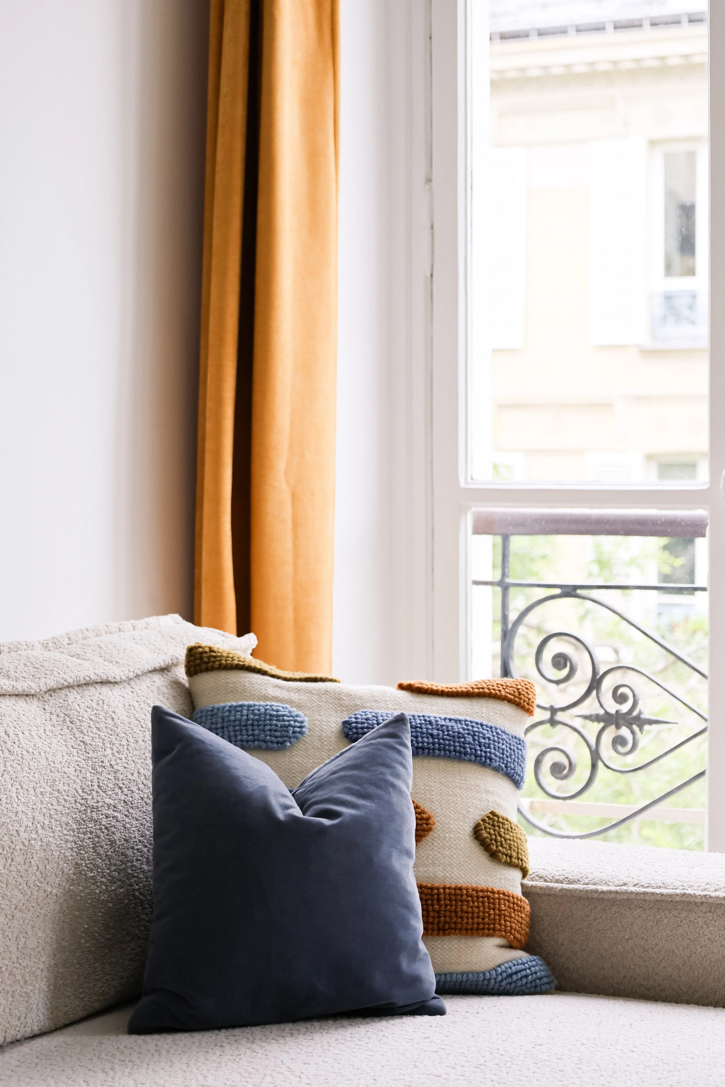 Coussin bleu en velours et coussin en laine avec motifs colorés sur canapé beige, près d'une fenêtre avec rideaux orange.