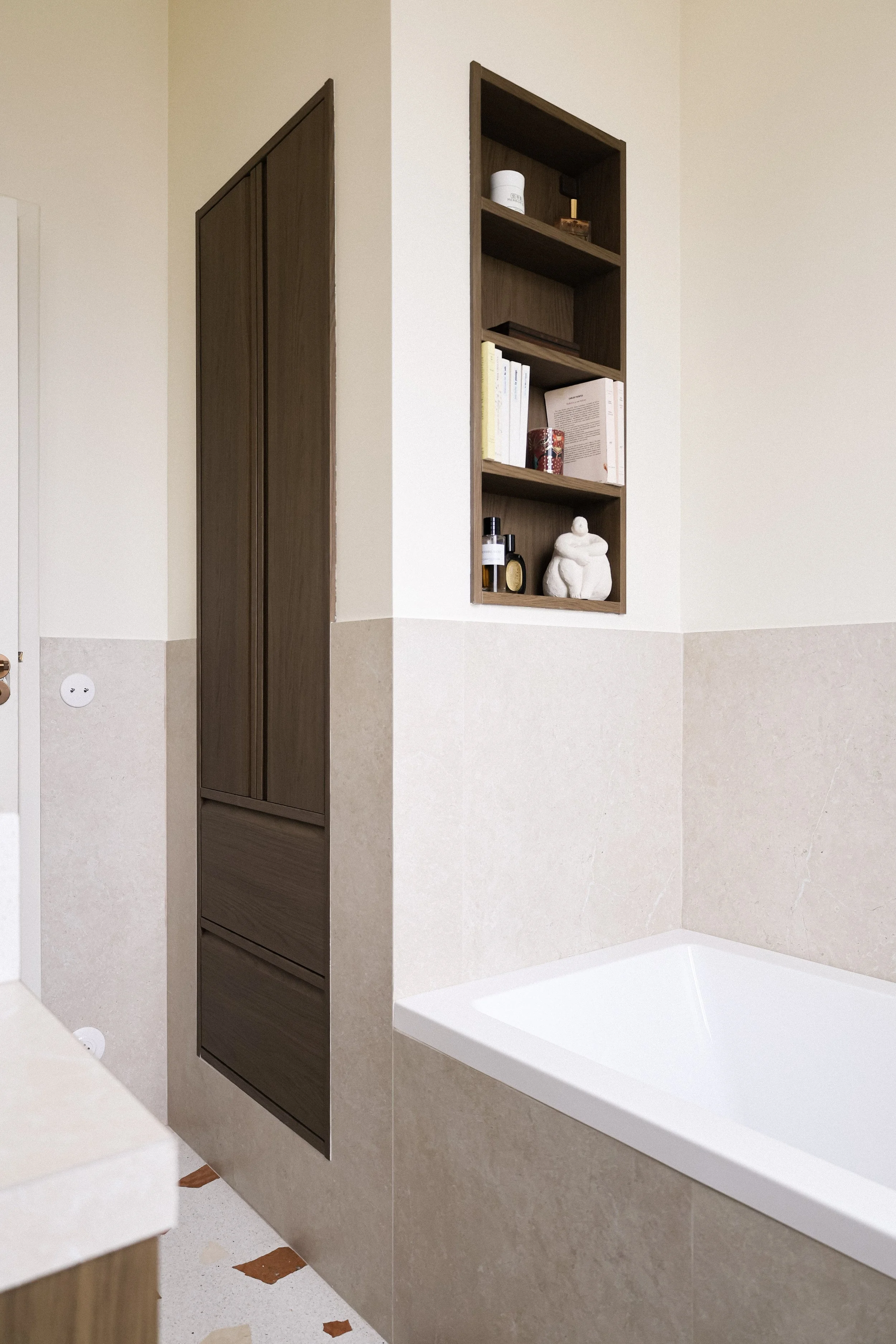 Une baignoire blanche encastrée dans un mur beige avec une étagère en bois au-dessus contenant des livres et des décorations.