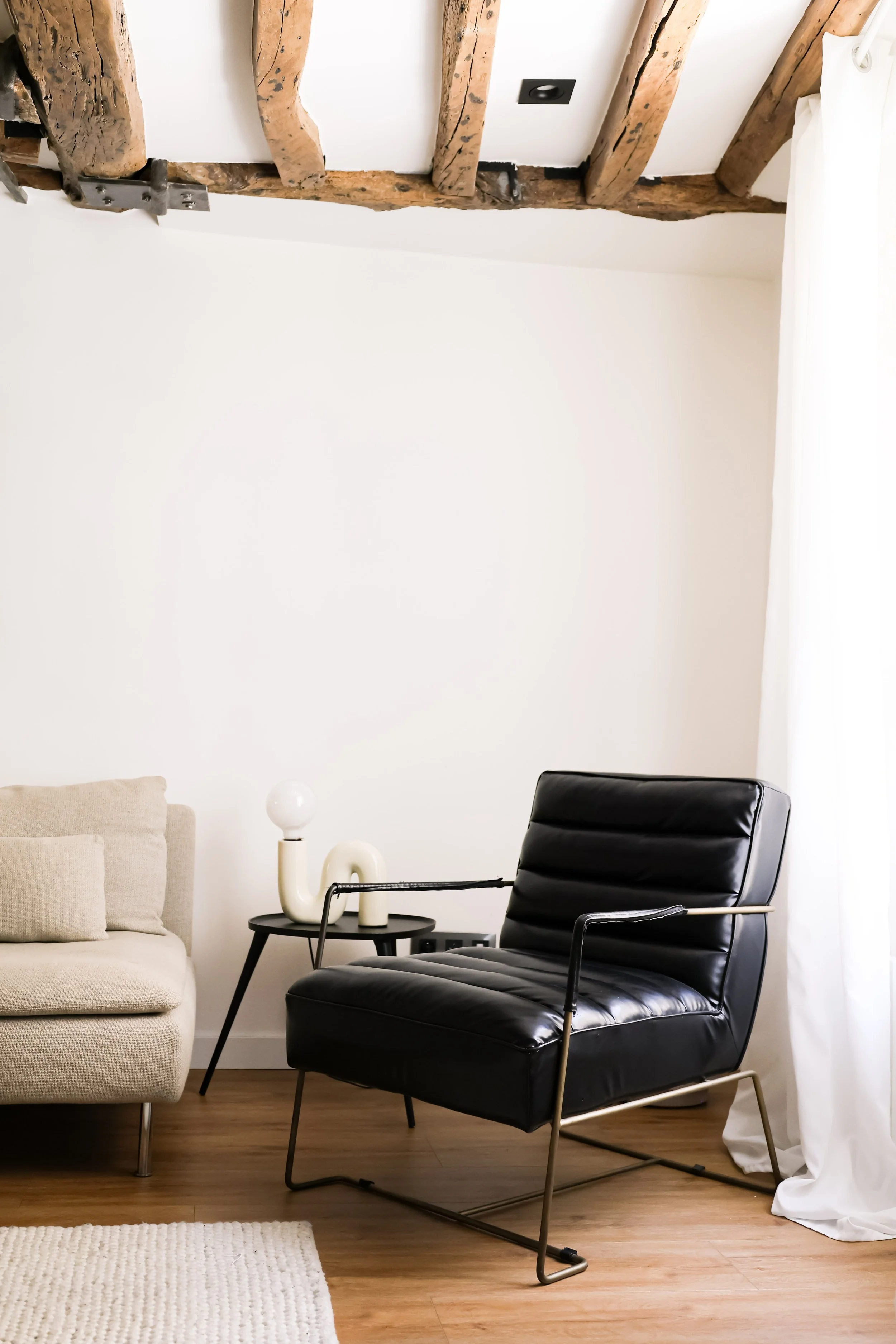 Un coin salon avec une banquette beige, une chaise en cuir noir avec un cadre en métal, une petite table noire avec une lampe et une décoration
