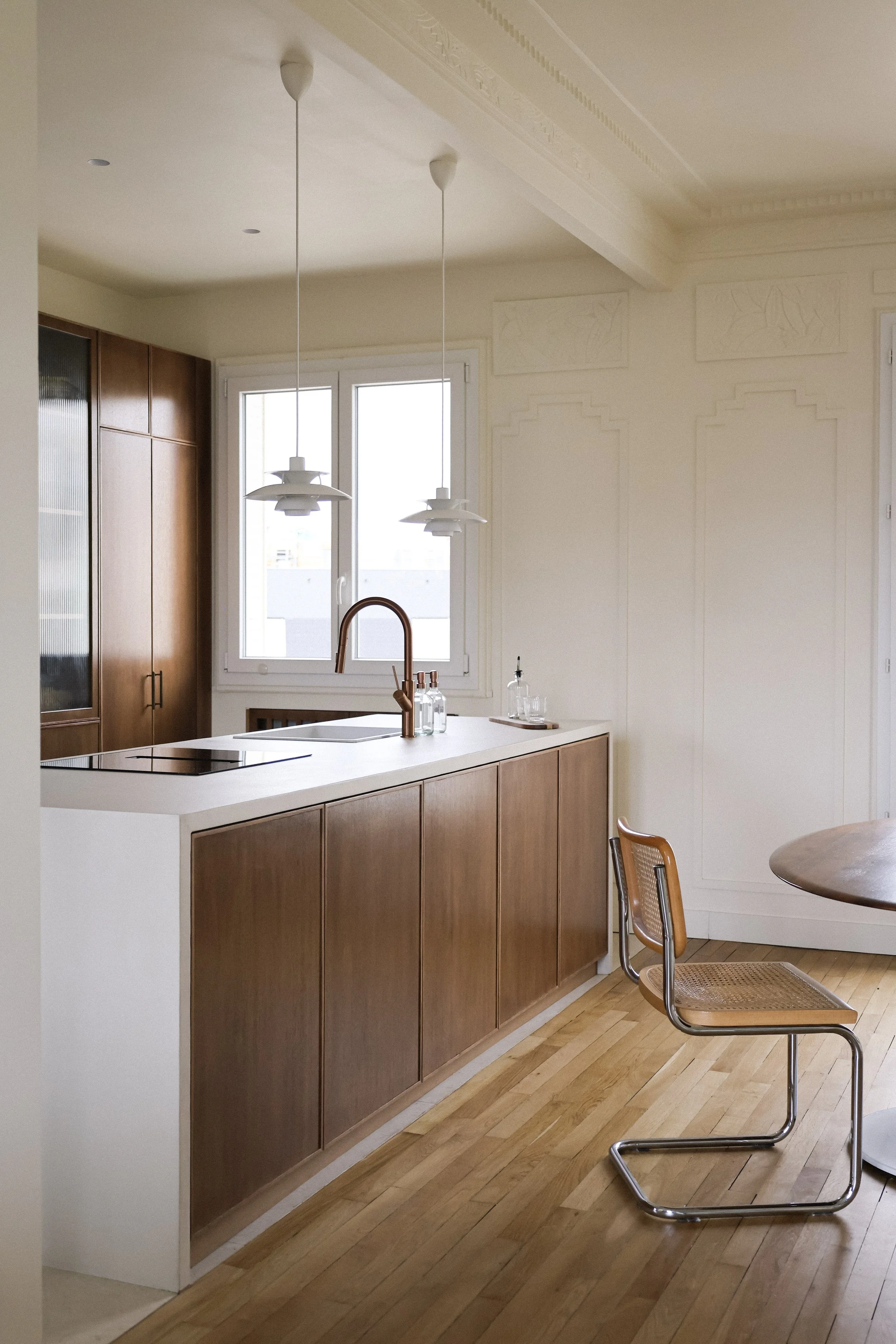Cuisine moderne avec plan de travail blanc, armoires en bois, deux lampes suspendues et une chaise. Fenêtre laissant entrer la lumière naturelle.