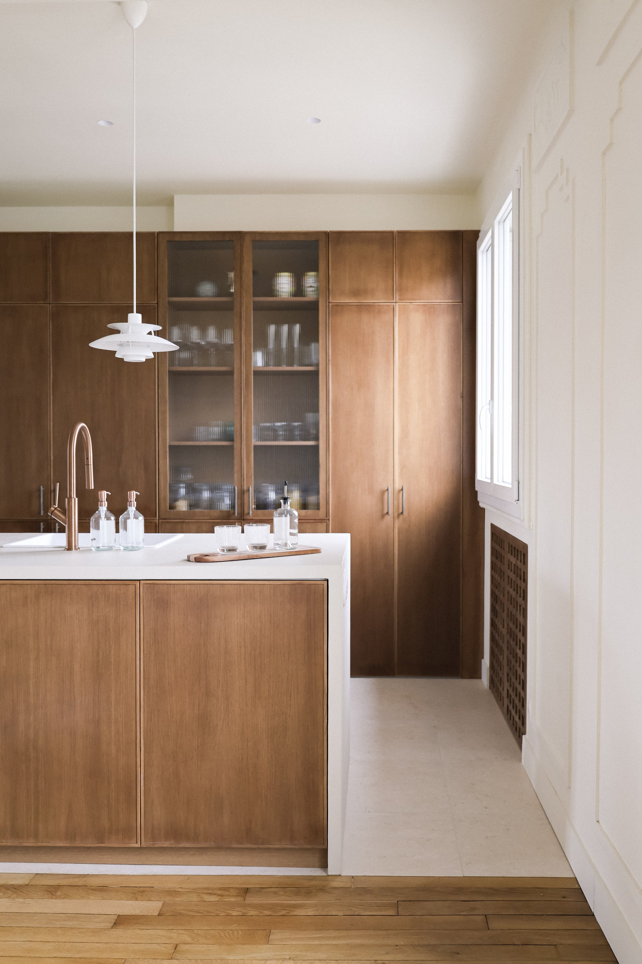 Cuisine moderne avec armoire en bois, plan de travail blanc, étagère avec vaisselle, fenêtre et luminaires suspendus blancs.