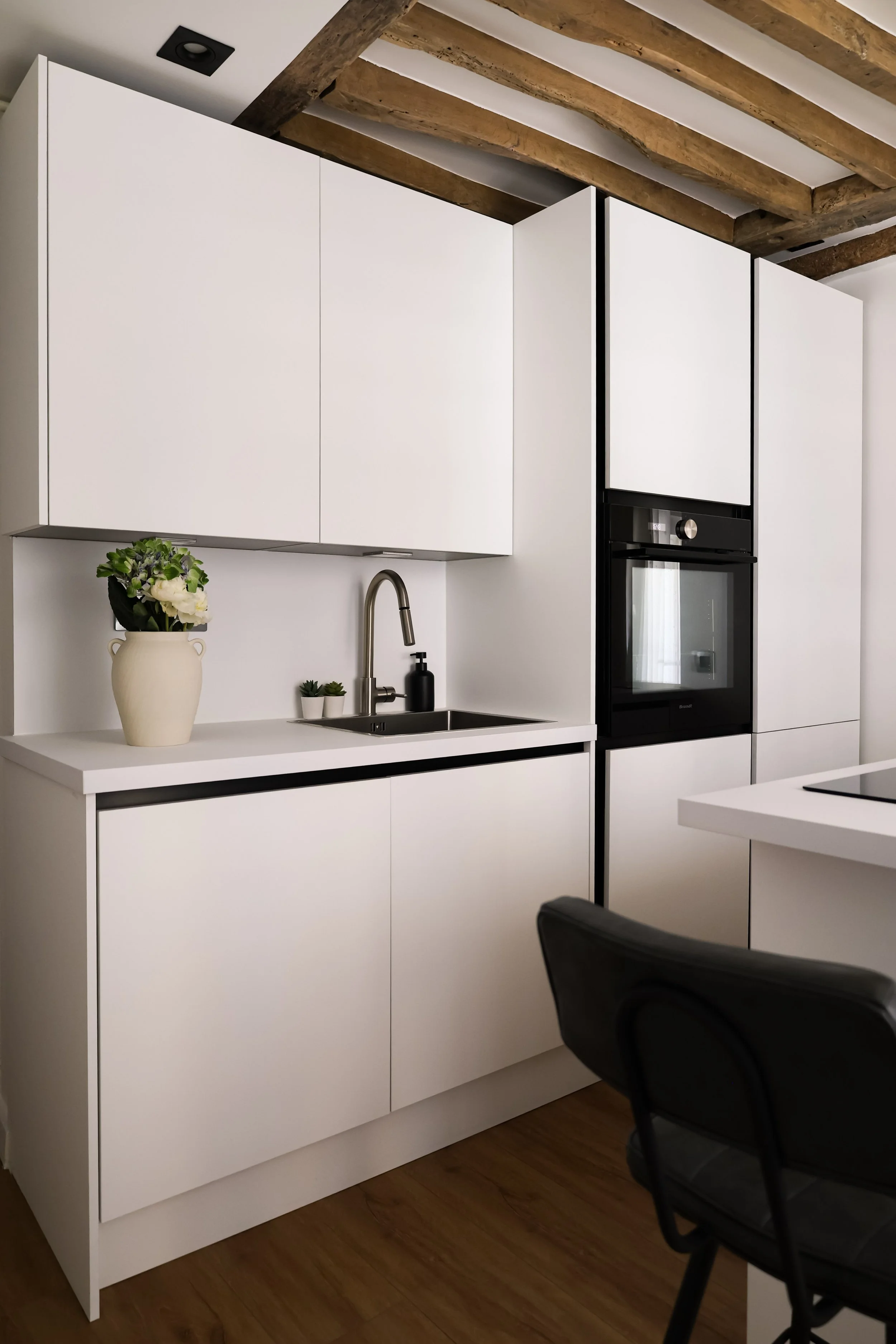 Cuisine moderne avec des armoires blanches, un évier en acier inoxydable, une plante en pot beige, un savon noir, un four encastré noir, un plafond avec poutres en bois, un plan de travail blanc, et une chaise noire.