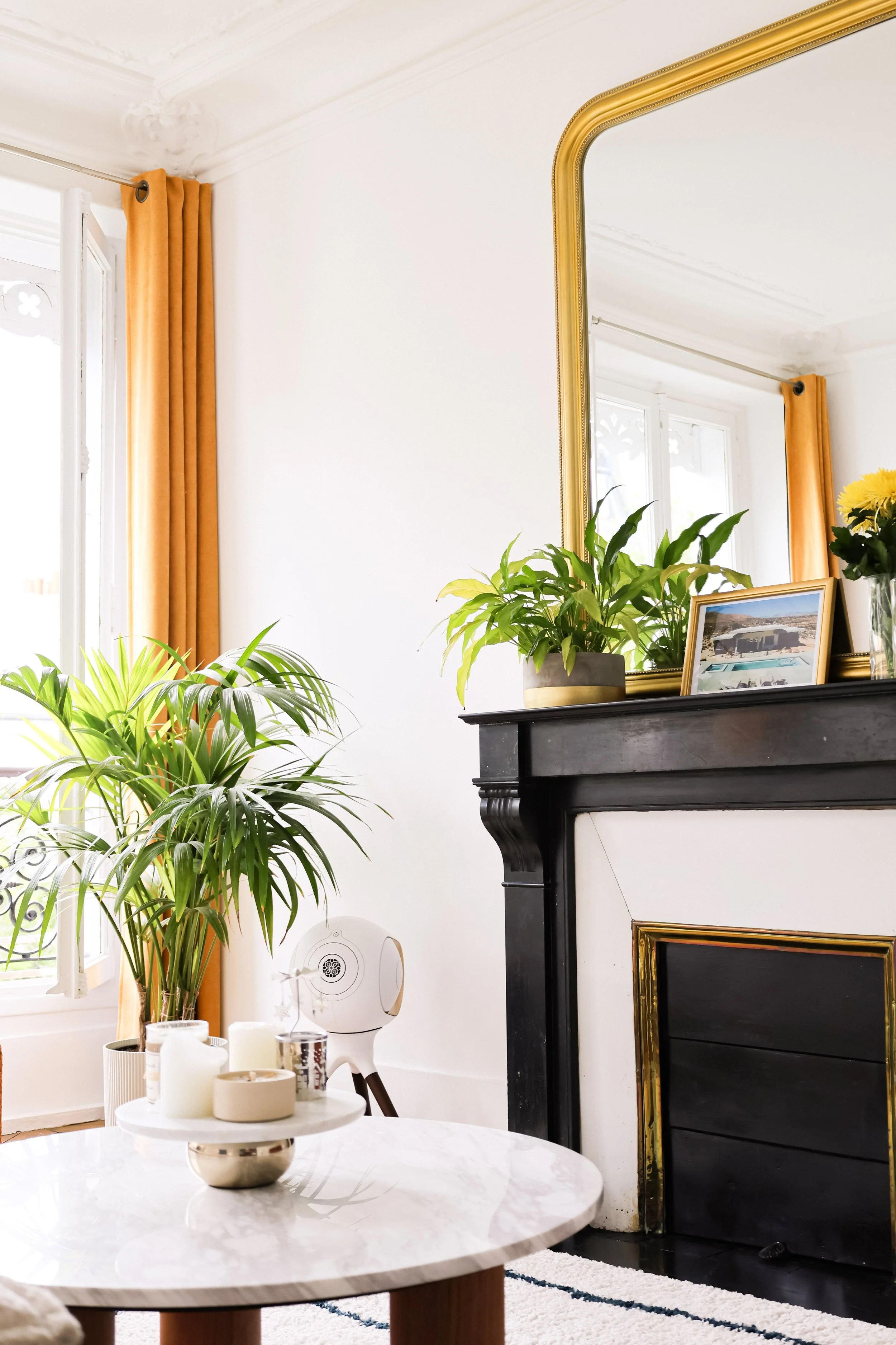 Salon lumineux avec cheminée noire, plantes vertes sur le manteau de cheminée, grand miroir doré, table basse en marbre avec bougies, gros coussin beige, et rideaux orange accentuant la décoration.
