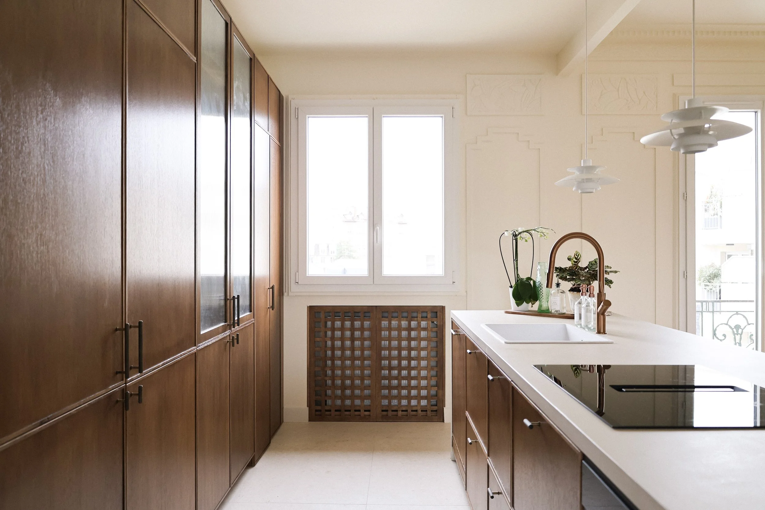 Cuisine moderne avec armoires en bois foncé, une fenêtre laissant entrer la lumière, un évier avec une robinetterie en cuivre, et un plan de travail en quartz clair décoré de plantes et de bouteilles.