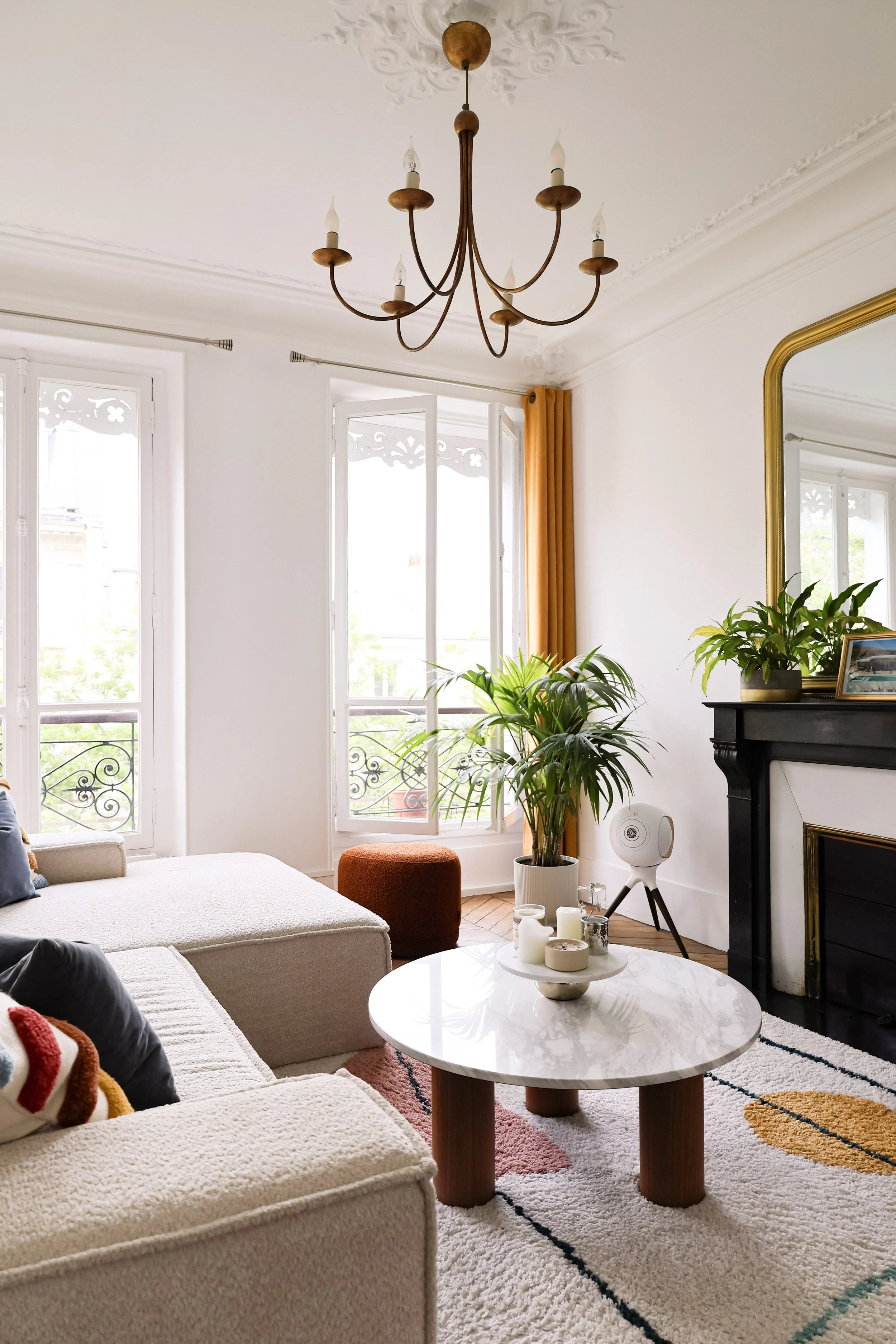 Salon lumineux avec canapés, table basse en marbre, cheminée noire, plantes vertes, miroir doré, chandeliers, et fenêtres ouvertes avec rideaux jaunes.