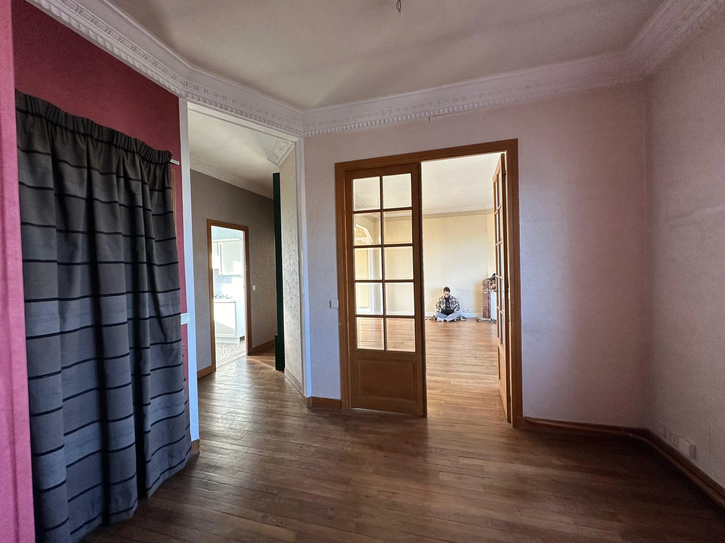 Intérieur d'une maison avec murs aux couleurs différentes, parquet en bois clair, une pièce visible à travers une porte vitrée en bois, une personne assise au sol dans cette pièce.