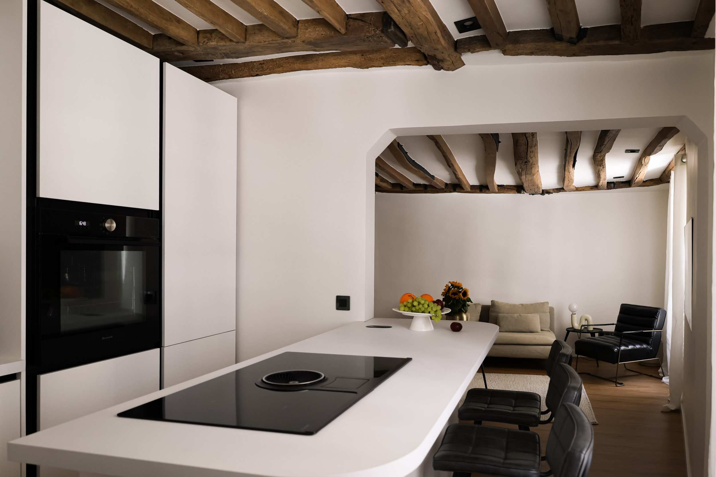 Intérieur d'une cuisine moderne avec un îlot blanc, des chaises noires, un mur blanc, des poutres apparentes en bois au plafond, et un coin salon avec canapé beige, fauteuil noir, table d'appoint et une décoration florale.