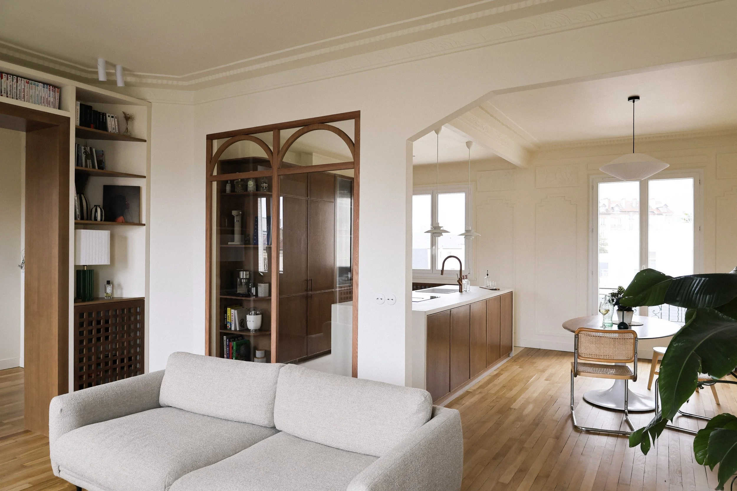 Intérieur d'un salon moderne avec canapé beige, bibliothèque en bois, espace cuisine avec plan de travail en bois, grande fenêtre, table ronde avec chaises, grande plante verte