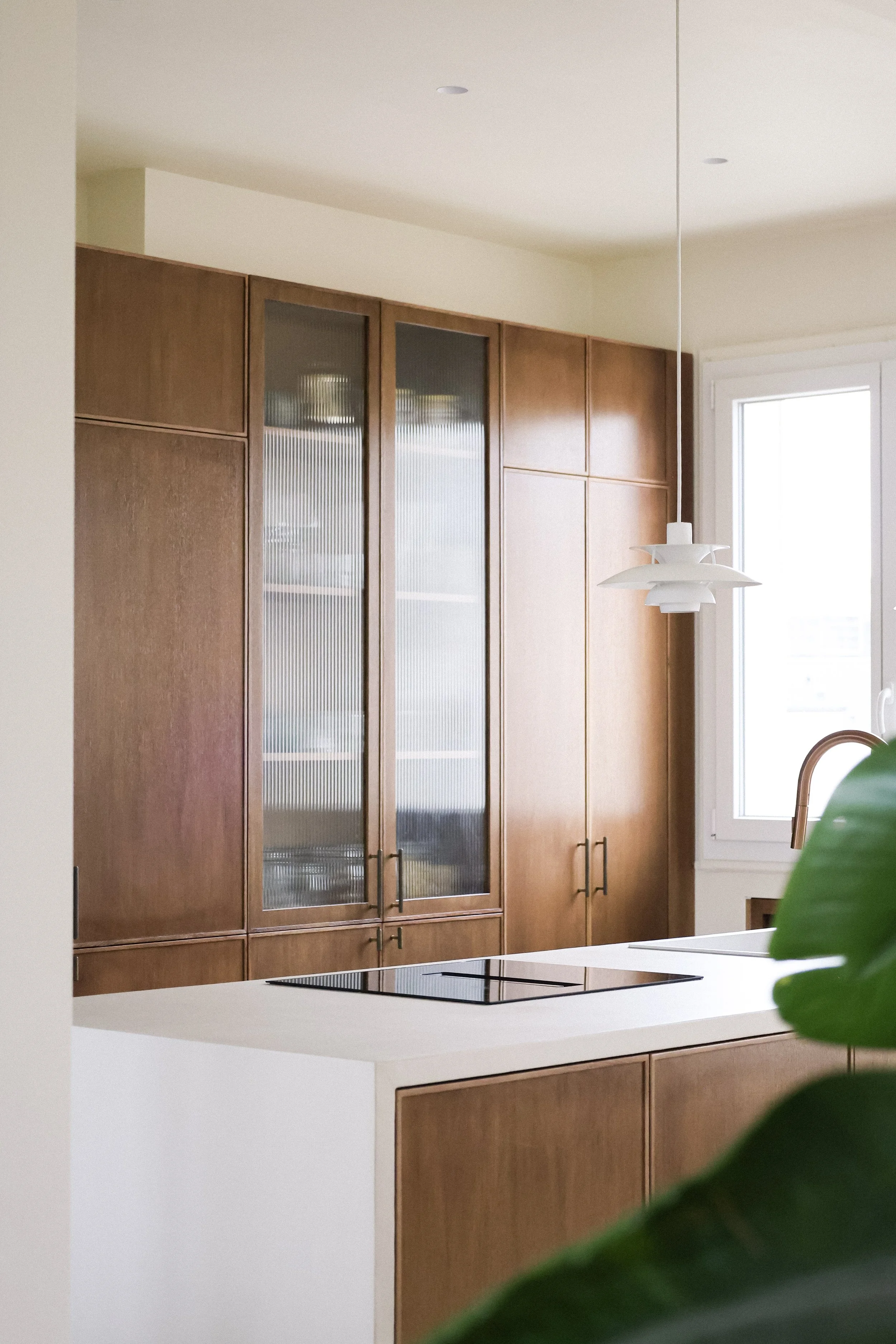 Cuisine moderne avec armoires en bois clair, plan de travail blanc, plaque de cuisson électrique intégrée, grande fenêtre, luminaire suspendu blanc, et plante verte en premier plan.