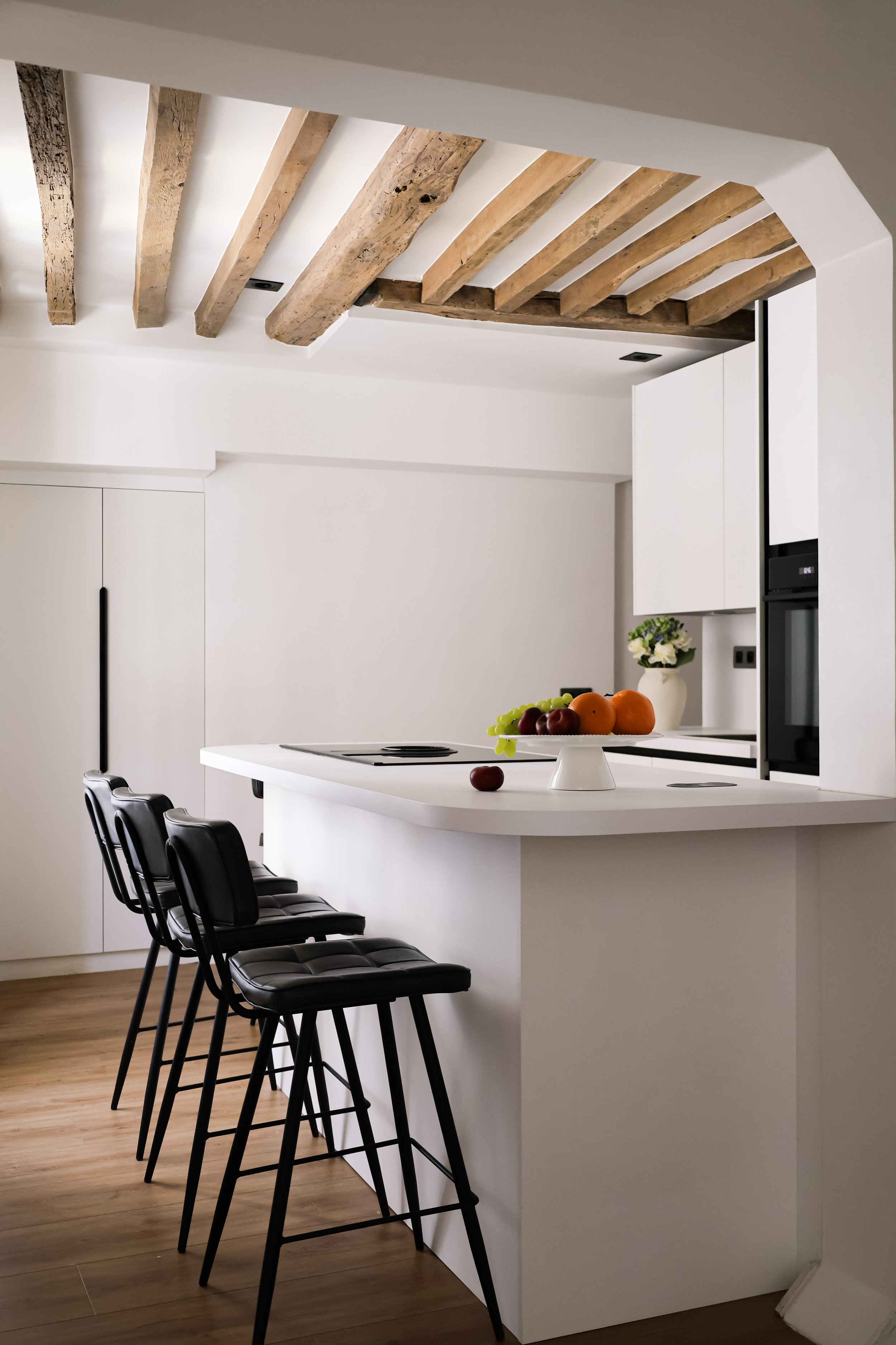 Cuisine moderne avec un plan de travail blanc, des chaises hautes noires en cuir, un plafond en poutres en bois et un mur blanc