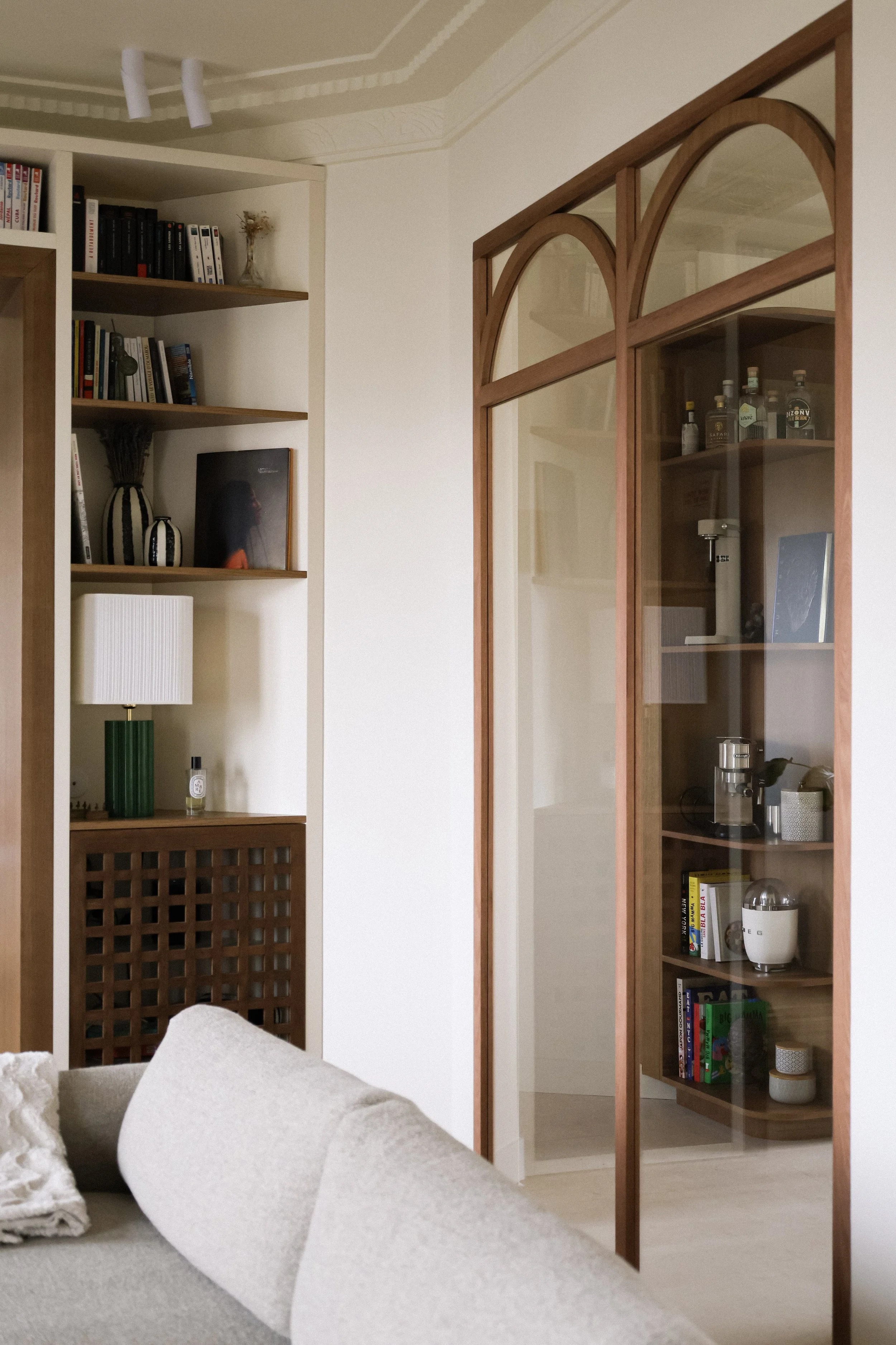 Intérieur d'une pièce moderne avec étagères en bois, une bibliothèque remplie de livres, un canapé gris, et un mur avec un mur en verre séparant une autre pièce contenant des bouteilles et des appareils électroménagers.