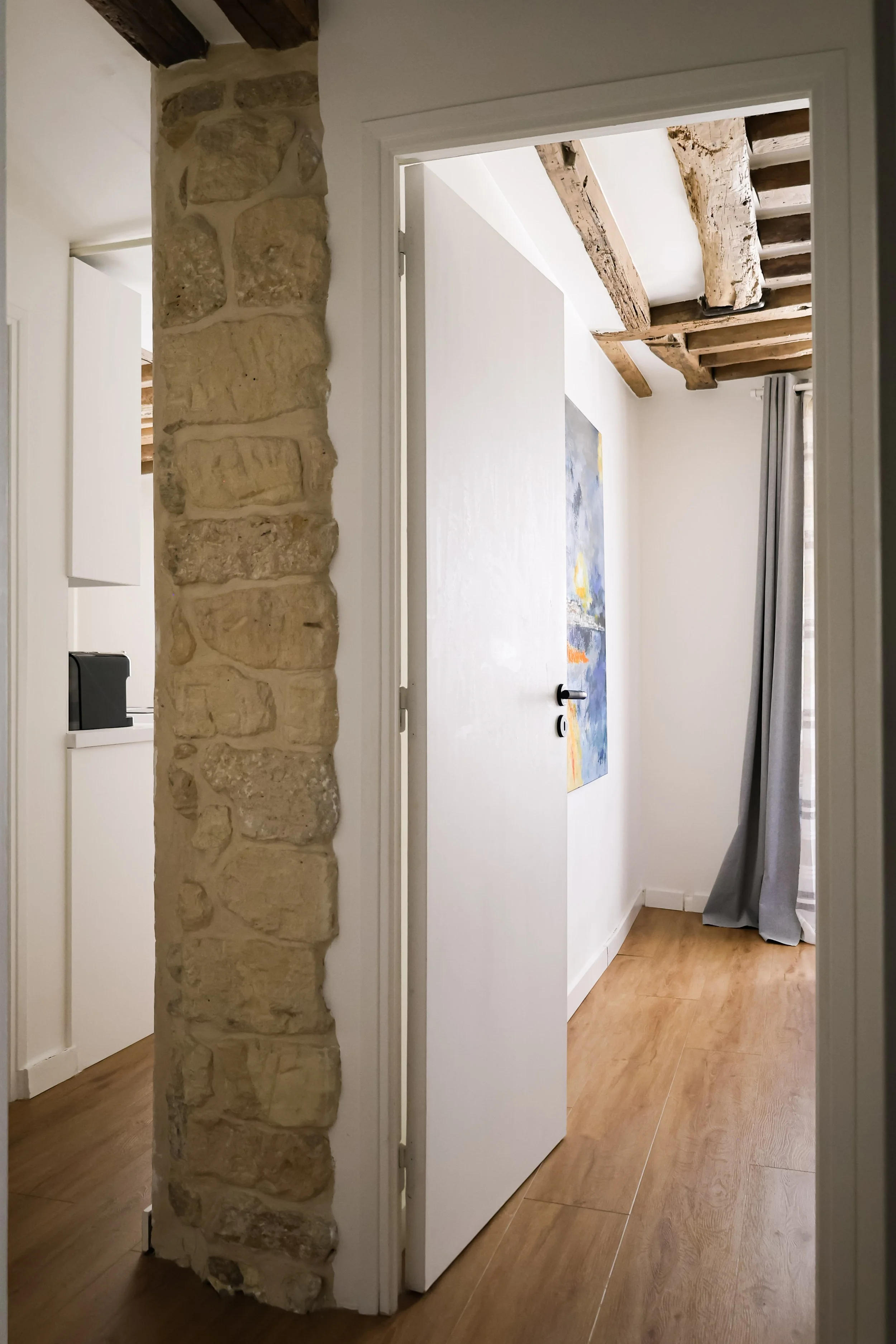 Une porte ouverte menant à une pièce avec un mur blanc, un tableau abstrait, un sol en bois clair, et un plafond avec des poutres en bois apparentes.